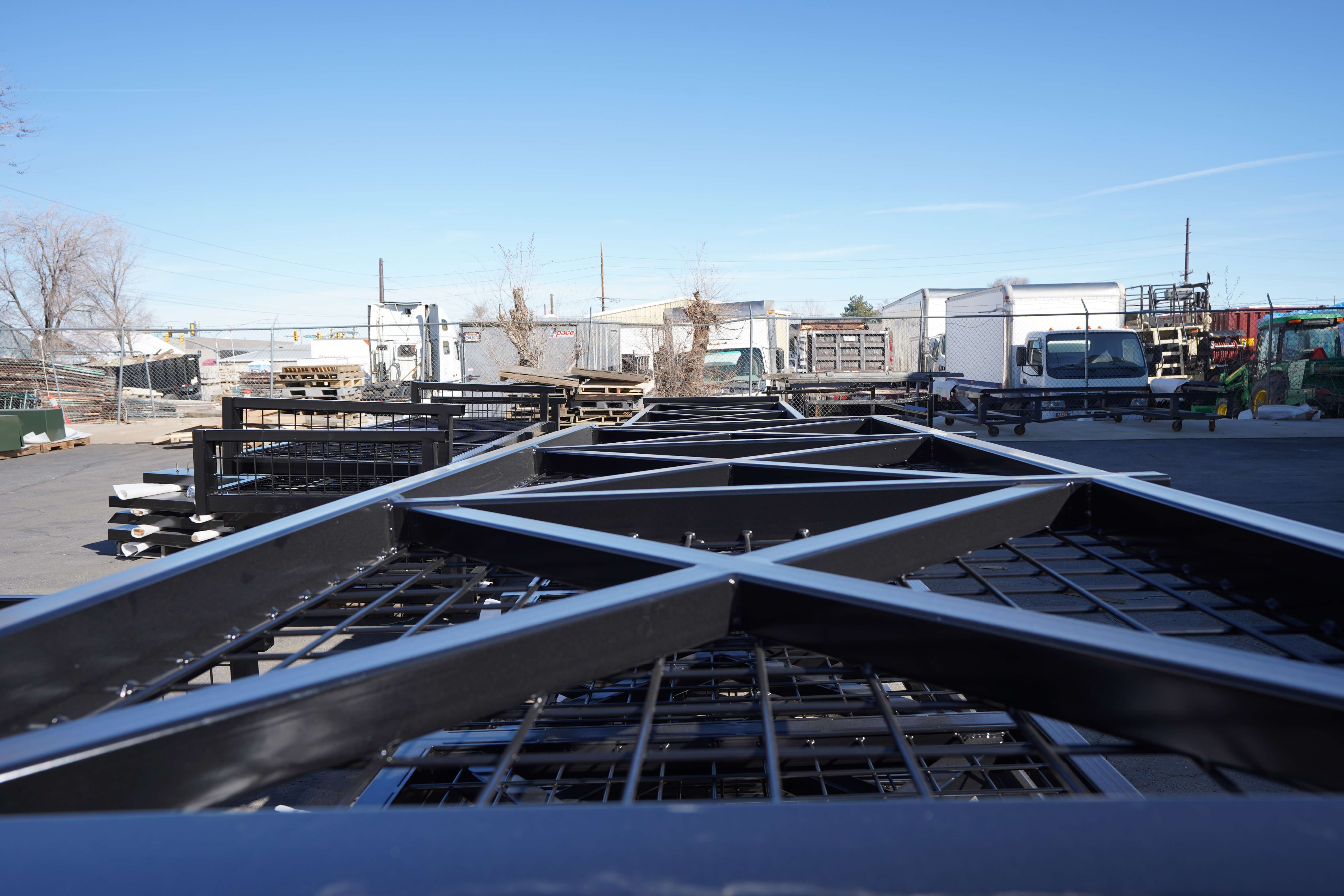 The image shows an outdoor yard of a metal fabrication facility. In the foreground, there are fabricated metal frames with grid or mesh panels that appear to be finished or partially finished metal railings or fence panels similar to those seen in previous images. The black metal structures have X-pattern bracing and grid infill panels.