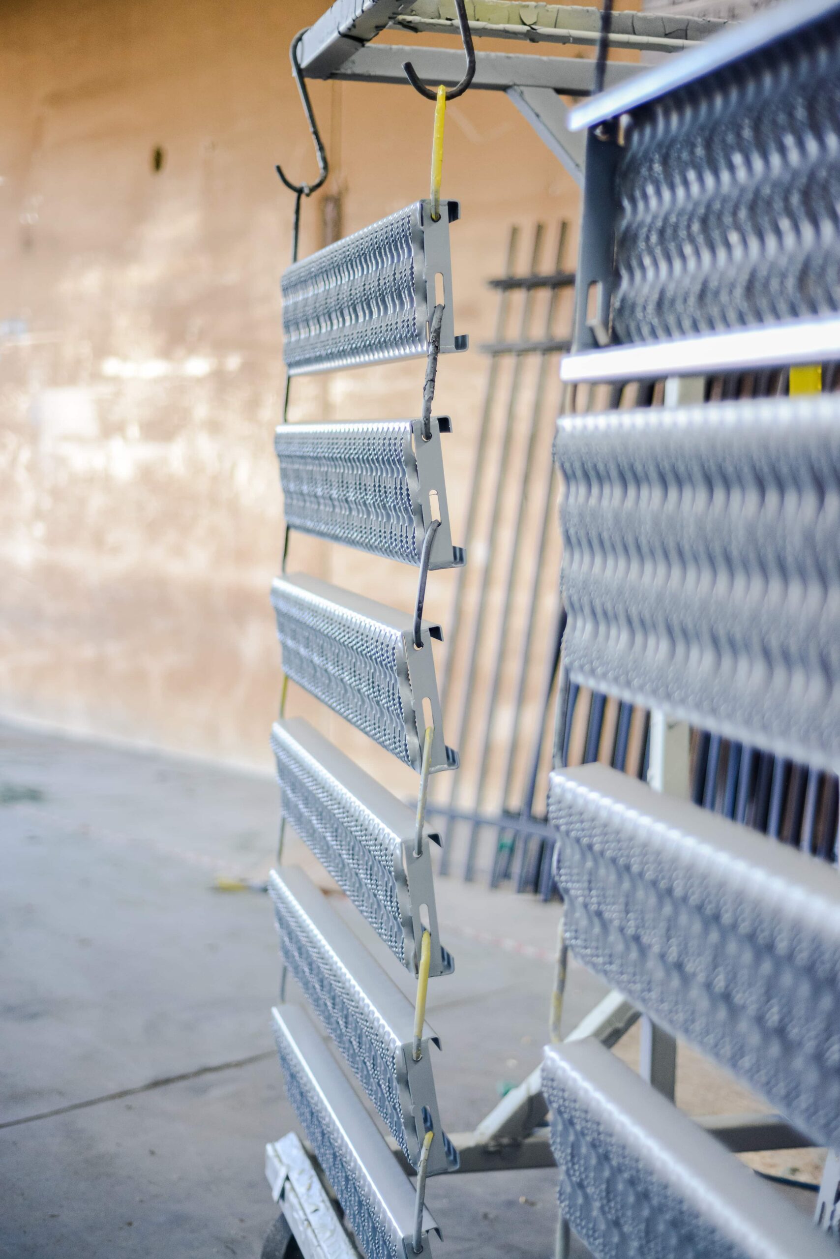 Multiple silver metal venetian blinds or louvered panels suspended from hooks in a powder coating facility. The horizontal slats have a metallic finish and are hung vertically in rows using yellow straps. These components appear to be freshly coated aluminum window blinds or similar architectural elements arranged for drying or curing in the industrial workspace. The facility's concrete floor and wall are visible in the background.
