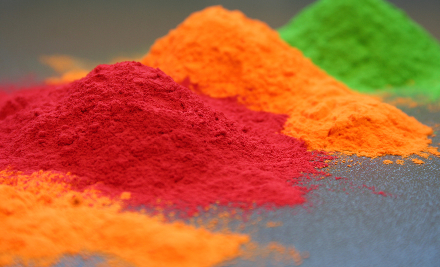 Close-up of vibrant powder coating pigments in red, orange, and green, piled in soft mounds on a neutral background, showcasing the texture and rich colors used in industrial finishing processes