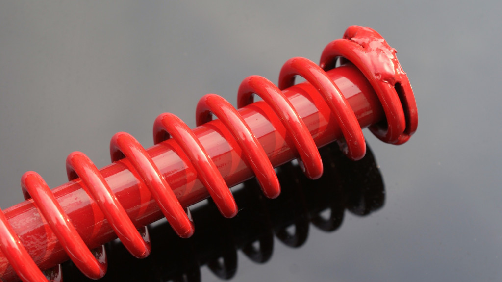 A close-up image of a bright red powder-coated metal spring. The image shows the distinctive helical coil structure of the spring with its vibrant glossy red finish, demonstrating the even and durable coating achieved through the powder coating process. The spring is photographed against a neutral gray background with a reflective surface that creates a shadow beneath it. This image highlights how powder coating can be applied to complex shapes like springs while providing a uniform, protective finish that enhances both the appearance and durability of metal components.