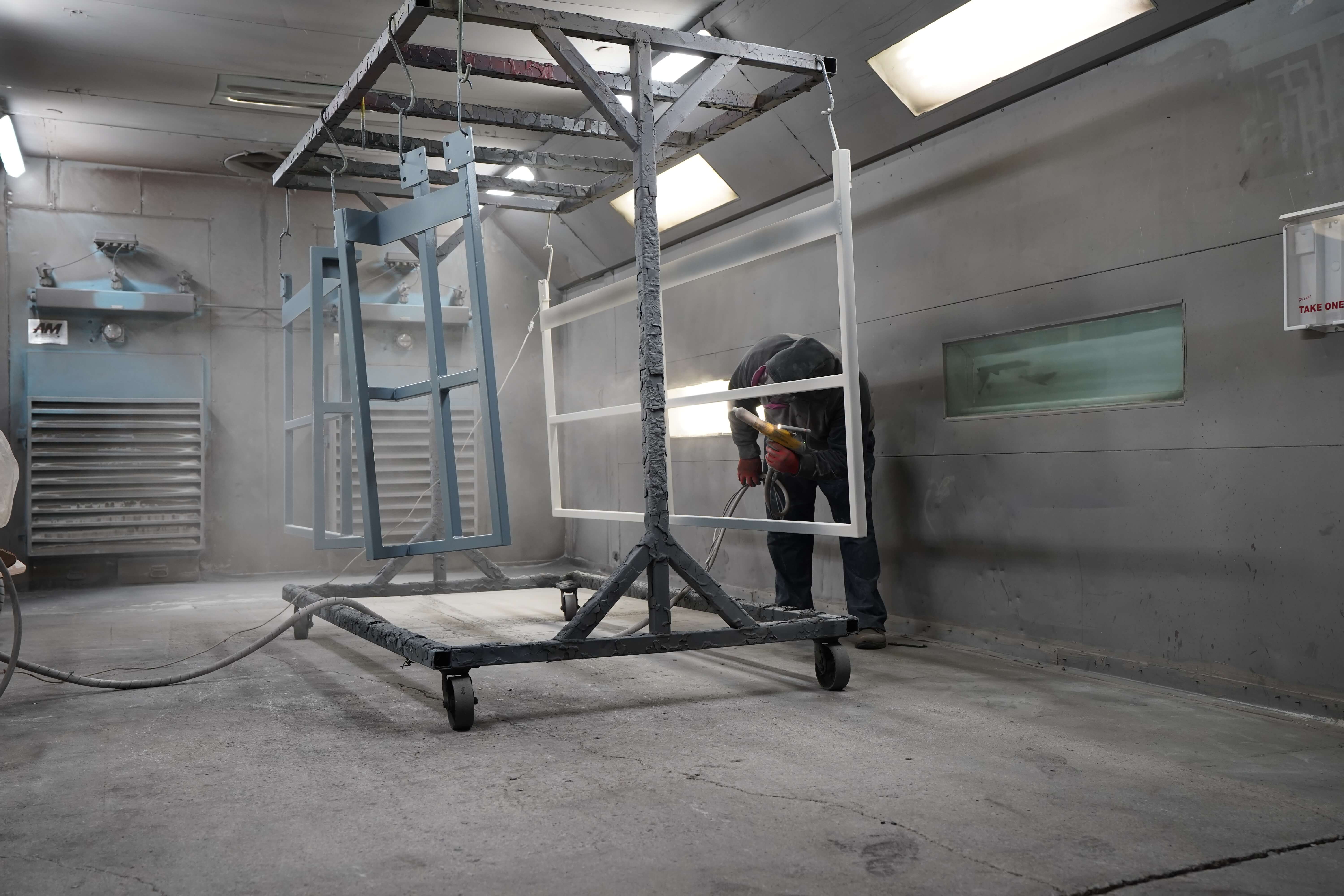 This image shows a powder coating spray booth or painting enclosure in an industrial facility. A worker wearing protective gear is operating a powder coating spray gun on the right side, applying powder to metal frames hanging from a wheeled rack system in the center of the booth. The booth features white walls and bright overhead lighting to provide good visibility during the coating process. Multiple metal parts are suspended from the rack - there appears to be a light blue frame and a white frame or panel being coated. The powder coating booth is equipped with ventilation systems visible on the left wall that help remove excess powder and maintain air quality. This represents a typical powder coating operation where metal parts are electrostatically sprayed before being transferred to a curing oven to create a durable finished surface.