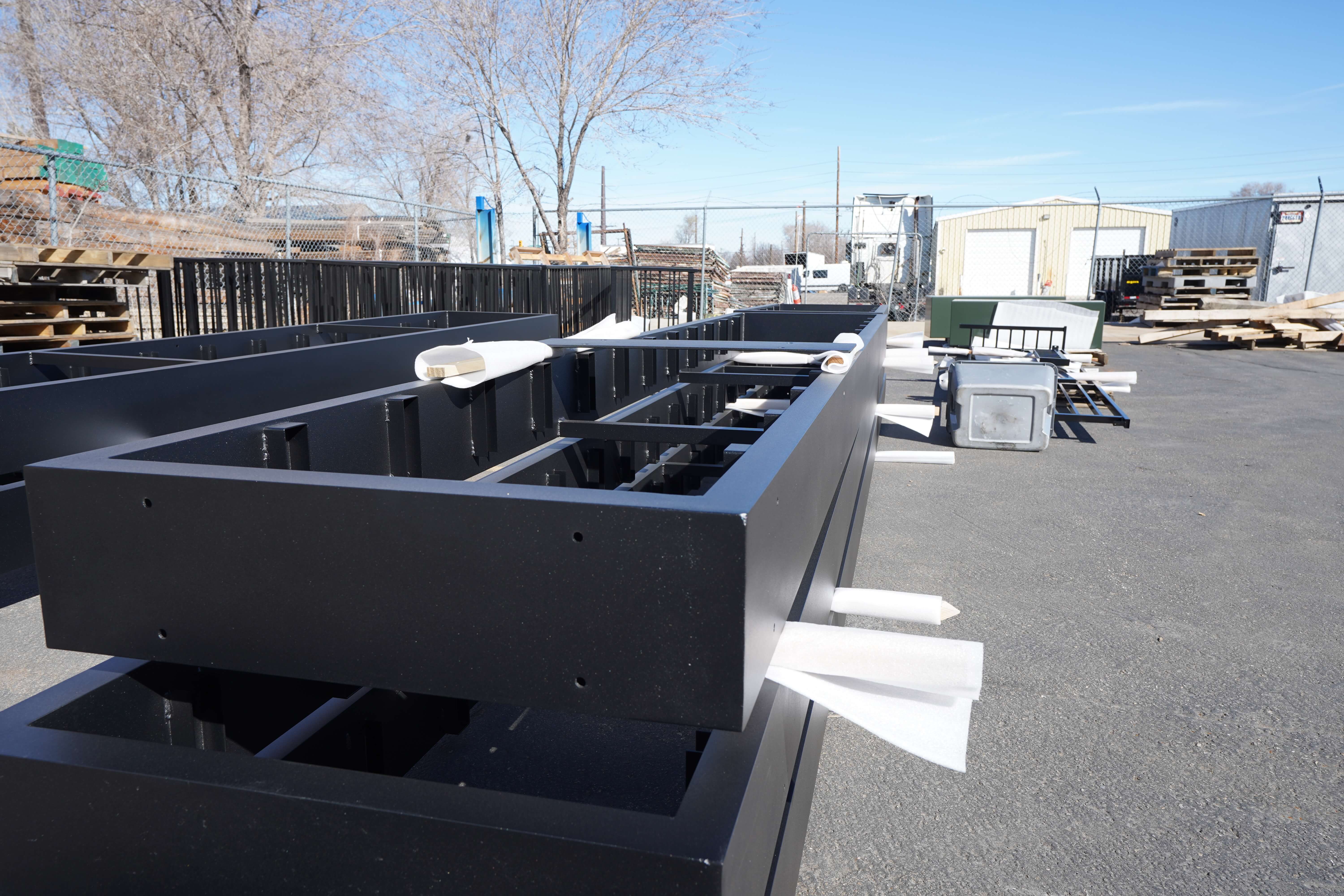 The black metal frames have a clean, finished appearance with protective white foam or plastic corner protectors attached to prevent damage during shipping or installation. The frames appear to be rectangular with vertical supports and are lined up on the asphalt surface of the yard