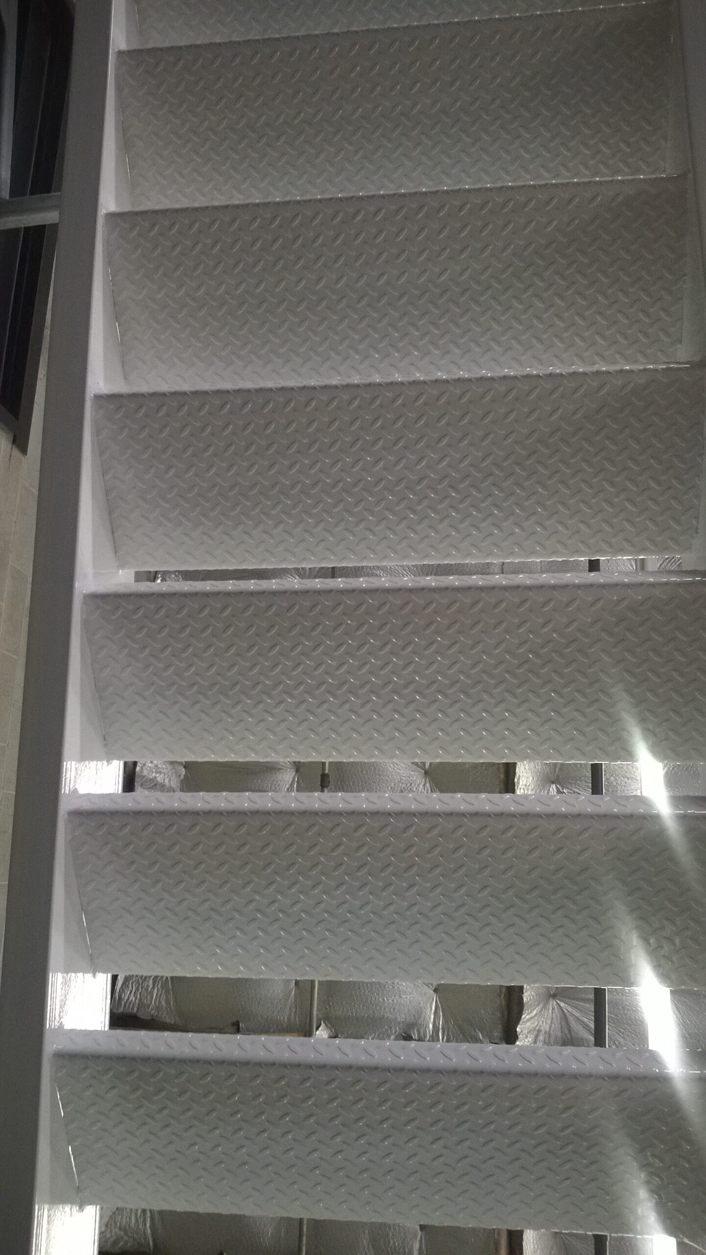 Close-up of industrial metal stairs with textured diamond plate steps, showing multiple levels with some steps wrapped in protective packaging, creating a geometric pattern of light and texture