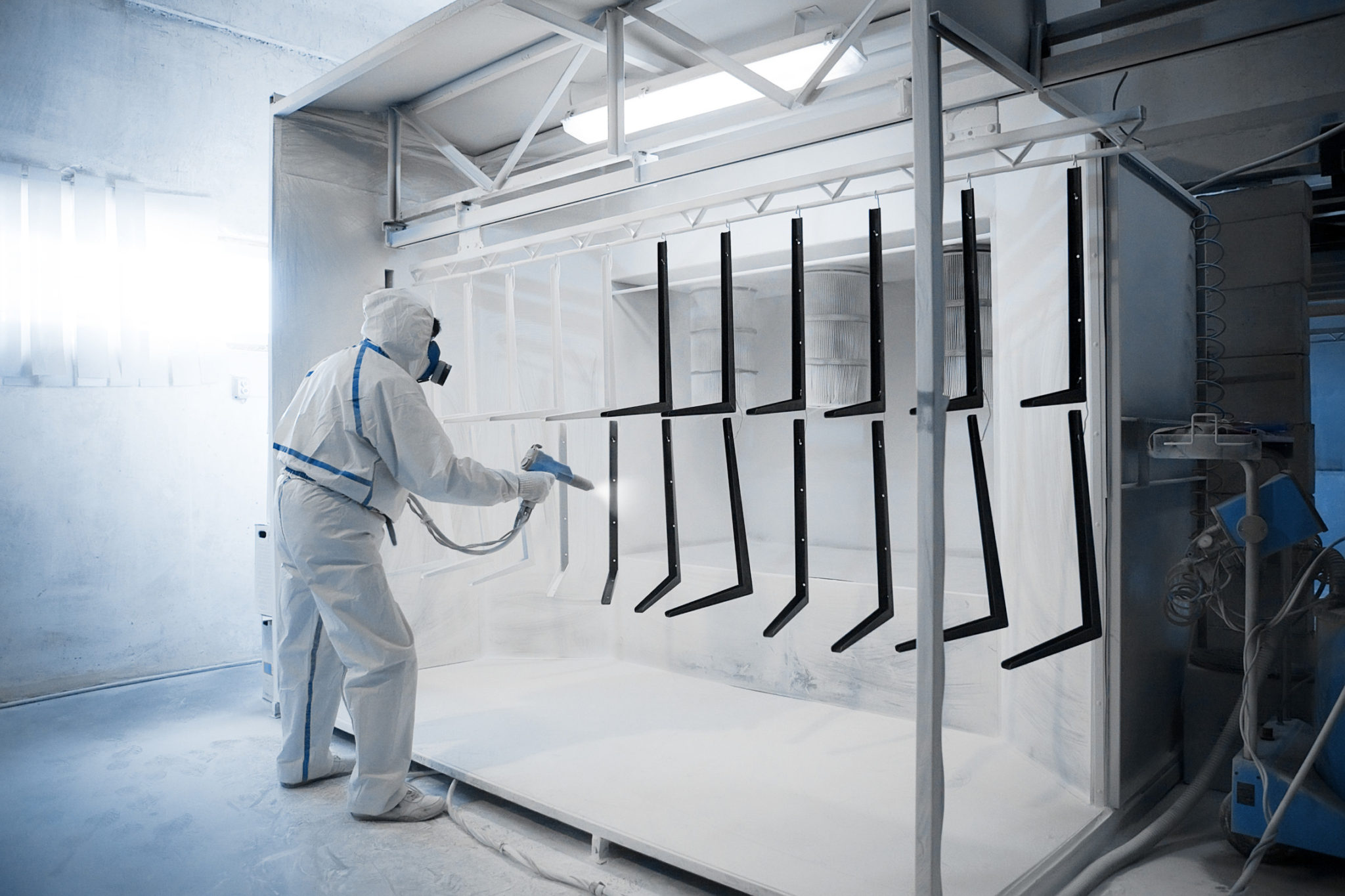 Worker in full protective white suit and respirator using a spray gun in a powder coating booth with hanging metal racks for industrial parts