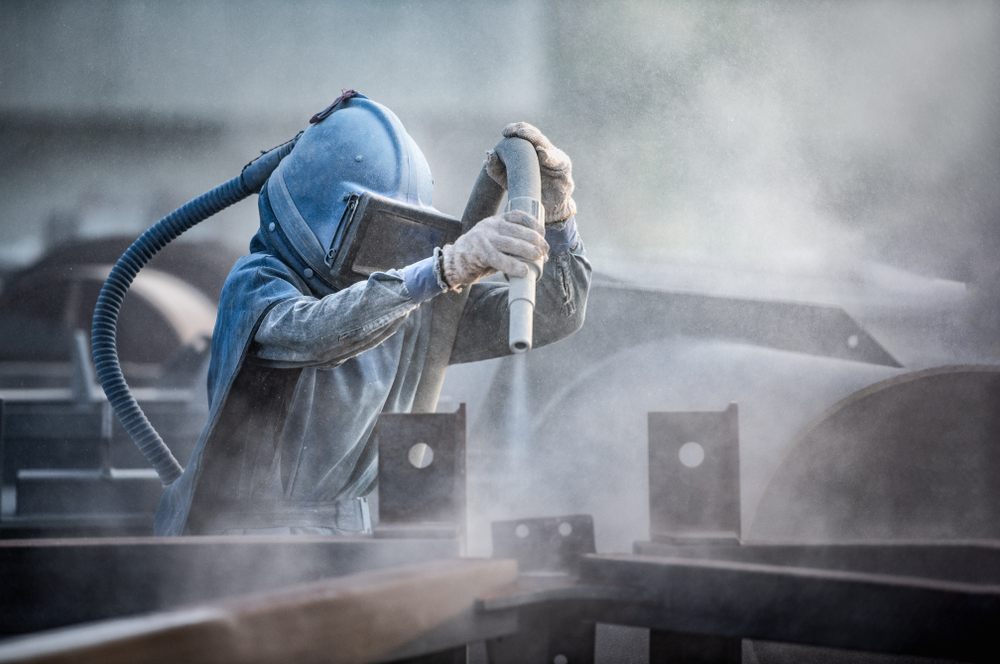 Industrial worker in blue protective suit and respirator mask performing sandblasting, surrounded by misty spray in a manufacturing environment