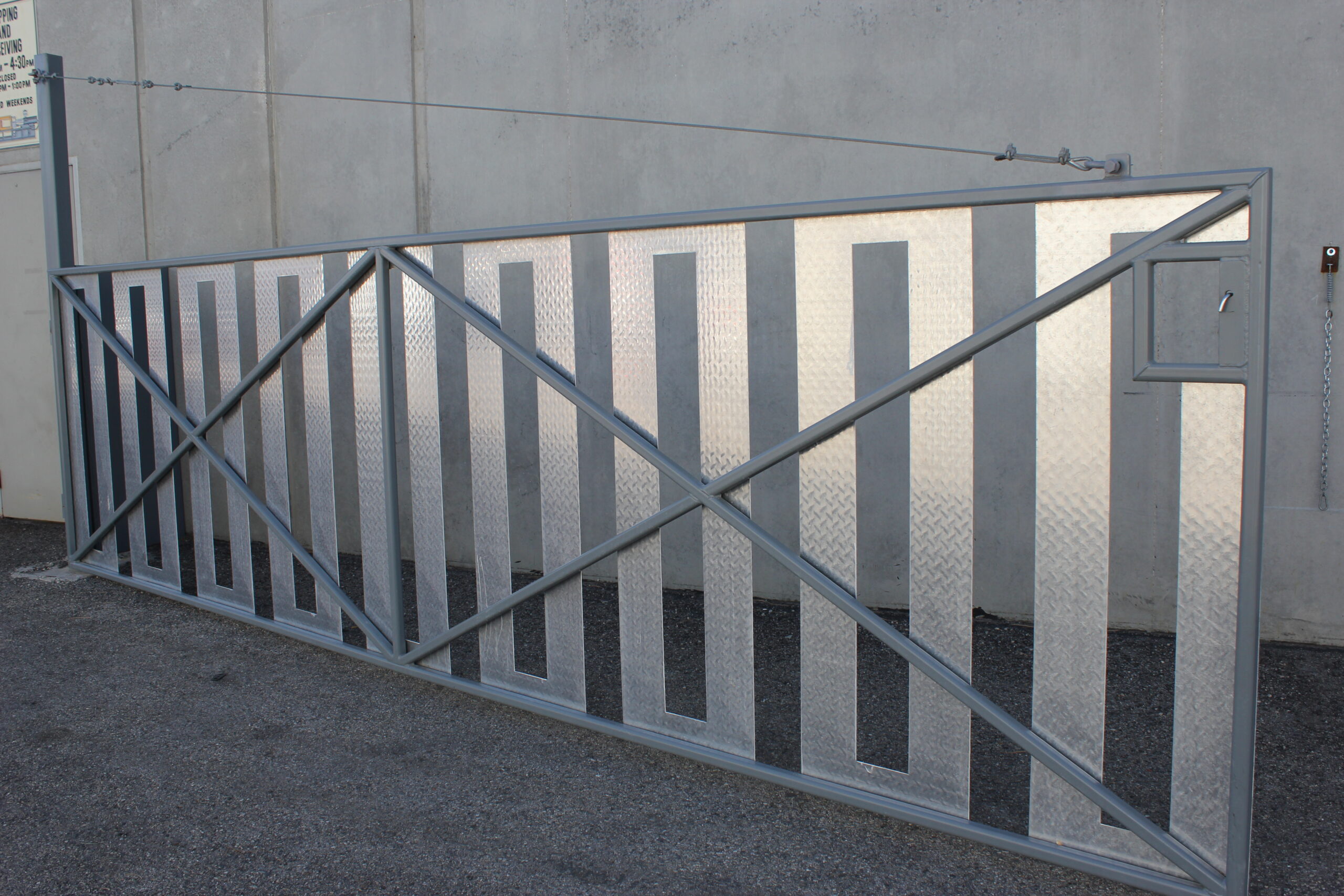 Industrial metal gate with crossbar design, featuring alternating gray and textured metal panels, mounted against a concrete wall with a textured asphalt ground, showcasing precision metalwork and industrial fabrication