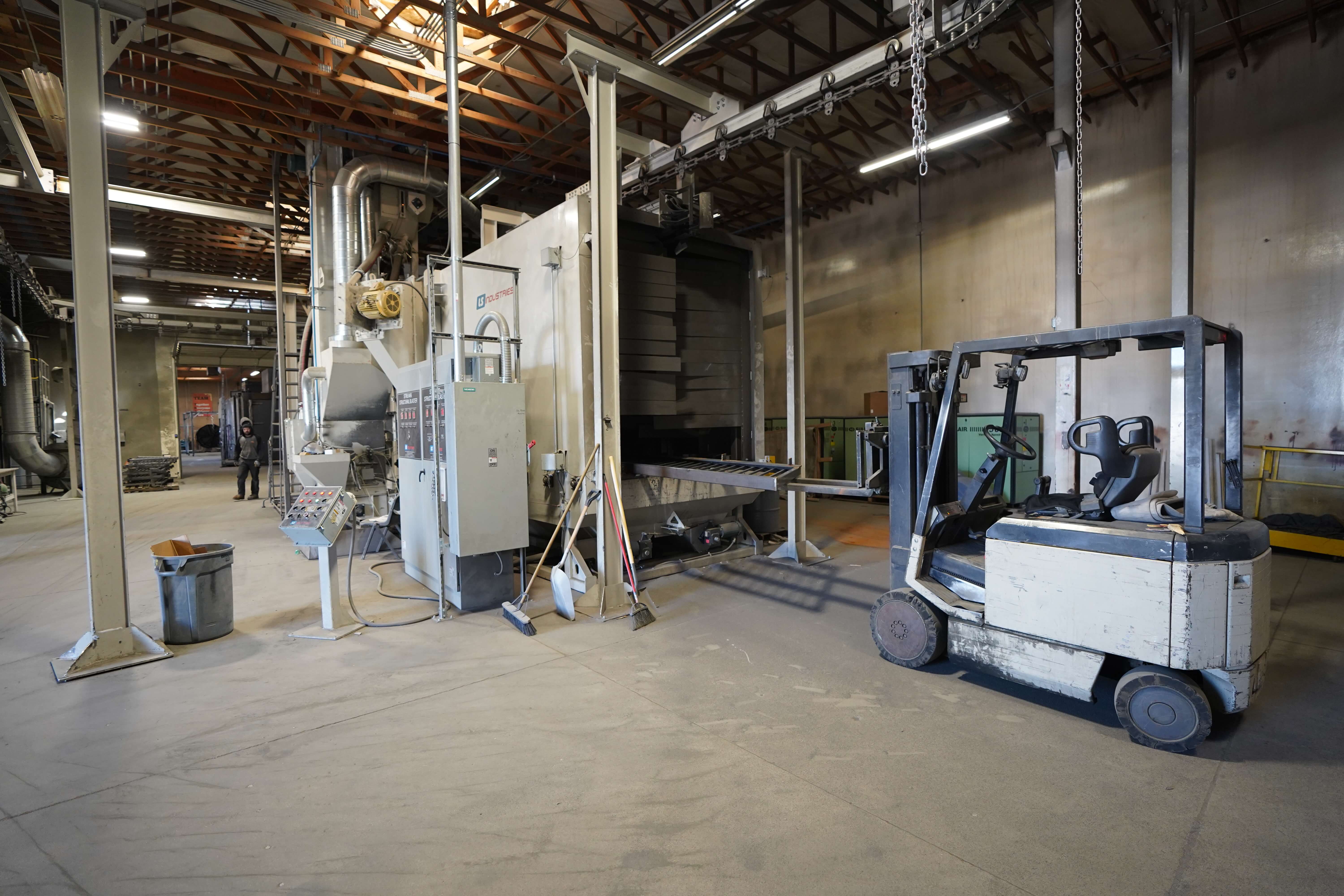 The image shows another area of the metal fabrication facility. This appears to be a powder coating or surface treatment section of the workshop. The space has a high ceiling with exposed wooden trusses and metal structural elements