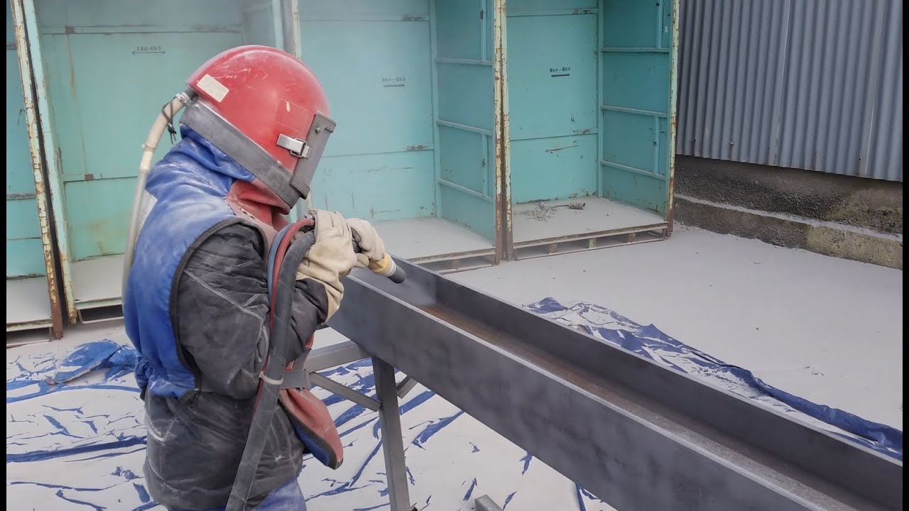Worker in blue protective jacket and red safety helmet performing sandblasting on a long metal beam, wearing protective gear in an industrial workshop with blue-green walls and protective floor covering