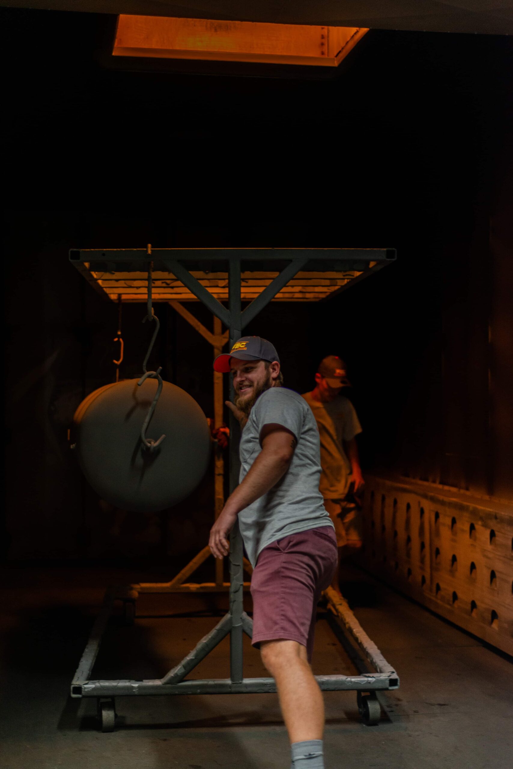 A worker in a powder coating facility pushing a metal rack cart through a curing oven. The employee wears a gray company cap, gray t-shirt and burgundy shorts while smiling at the camera. The cart holds a circular metal object and is moving through the orange-lit industrial oven with perforated walls. The darkened environment is illuminated primarily by the heat from the oven, creating an amber glow throughout the space. A second worker can be seen in the background.