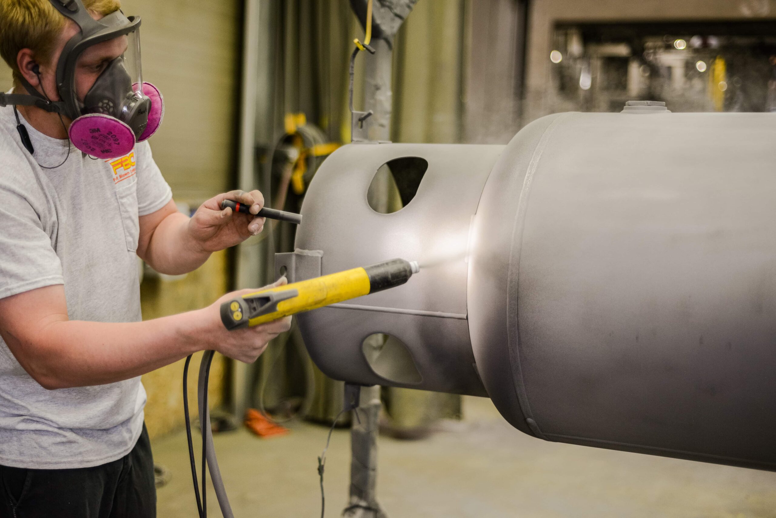 Worker applying powder coating to a large gray automotive or industrial part. The technician wears a respirator with pink filter cartridges and a white company t-shirt with yellow logo. He's using a yellow powder coating spray gun to apply a fine powder to the curved metallic component. The part appears to be a bumper or similar automotive element suspended in a spray booth, demonstrating the powder coating application process.
