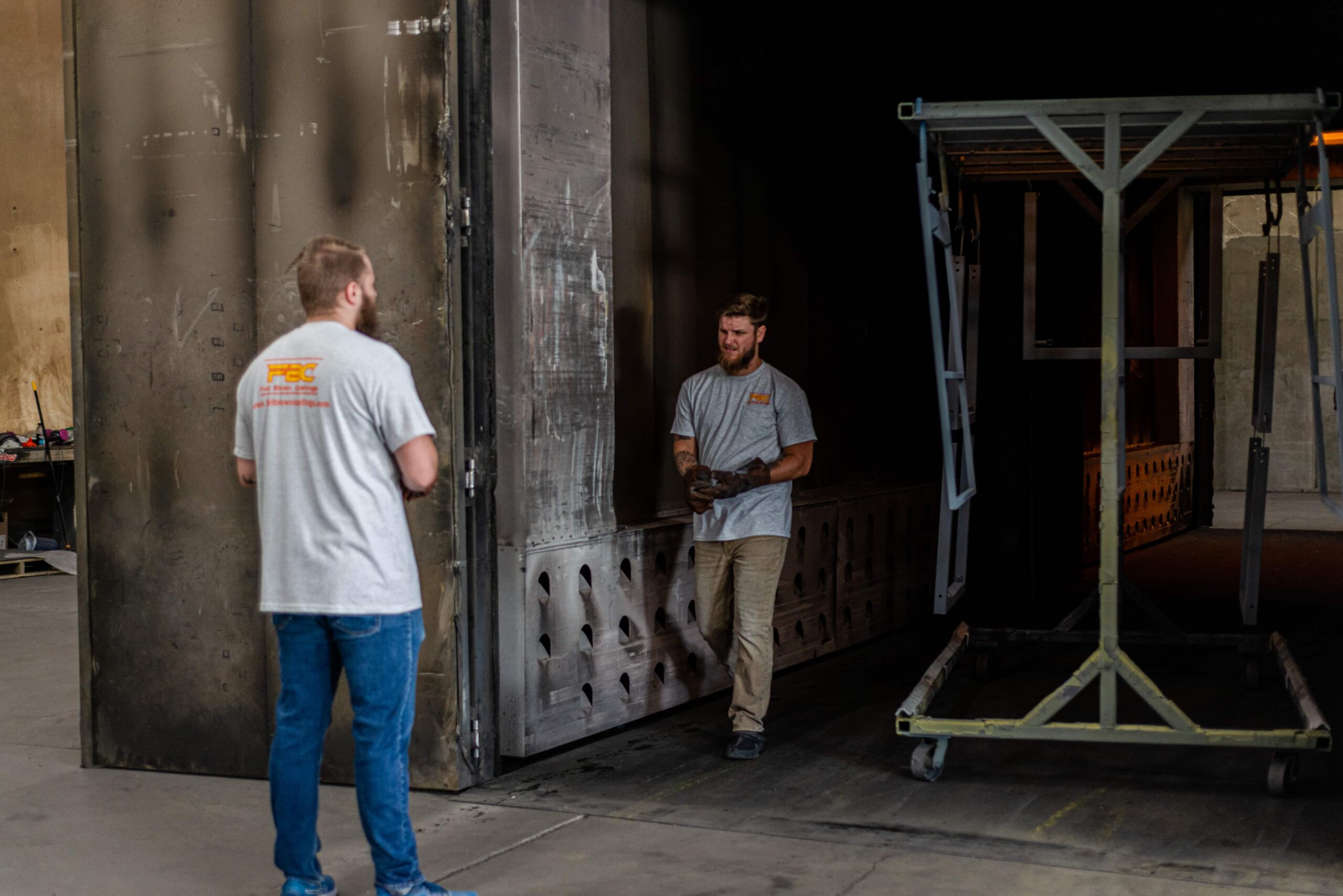 Two powder coating technicians in matching gray company shirts with an orange logo at a manufacturing facility. One worker stands outside the large industrial curing oven while another technician wearing work gloves is positioned inside the darkened oven chamber. A metal rack cart with blue framework is visible to the right of the oven entrance, used for transporting items through the powder coating process.