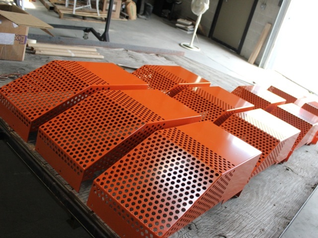 This image shows bright orange perforated metal panels or sheets that have been powder coated. The panels feature numerous evenly-spaced circular holes throughout their surface, creating a perforated pattern. They appear to be freshly coated or finished industrial components, arranged in rows on what looks like a wooden table or workbench in a manufacturing facility. The vibrant orange powder coating gives them a smooth, uniform finish. These types of perforated metal panels are commonly used in various applications such as machinery guards, ventilation covers, acoustic panels, or decorative architectural elements. The workshop environment is visible in the background with some tools and materials stored around the space.
