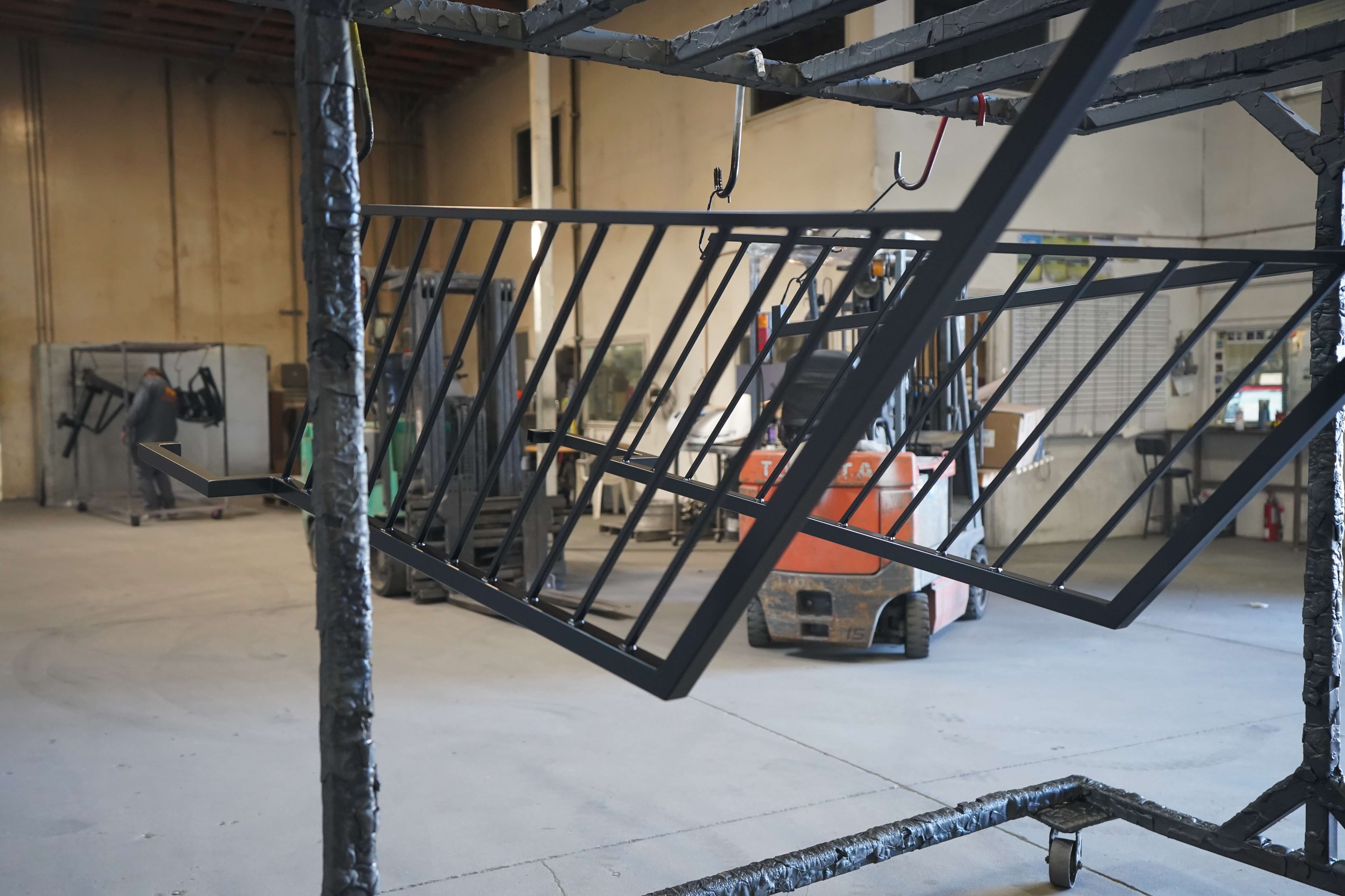 The image shows a metal fabrication workshop or manufacturing facility. In the foreground, there's a black metal railing or fence panel with vertical bars, partially assembled and appearing to be part of a larger structure under construction. The panel is supported by a welded metal frame structure with hooks visible at the top for hanging or mounting.