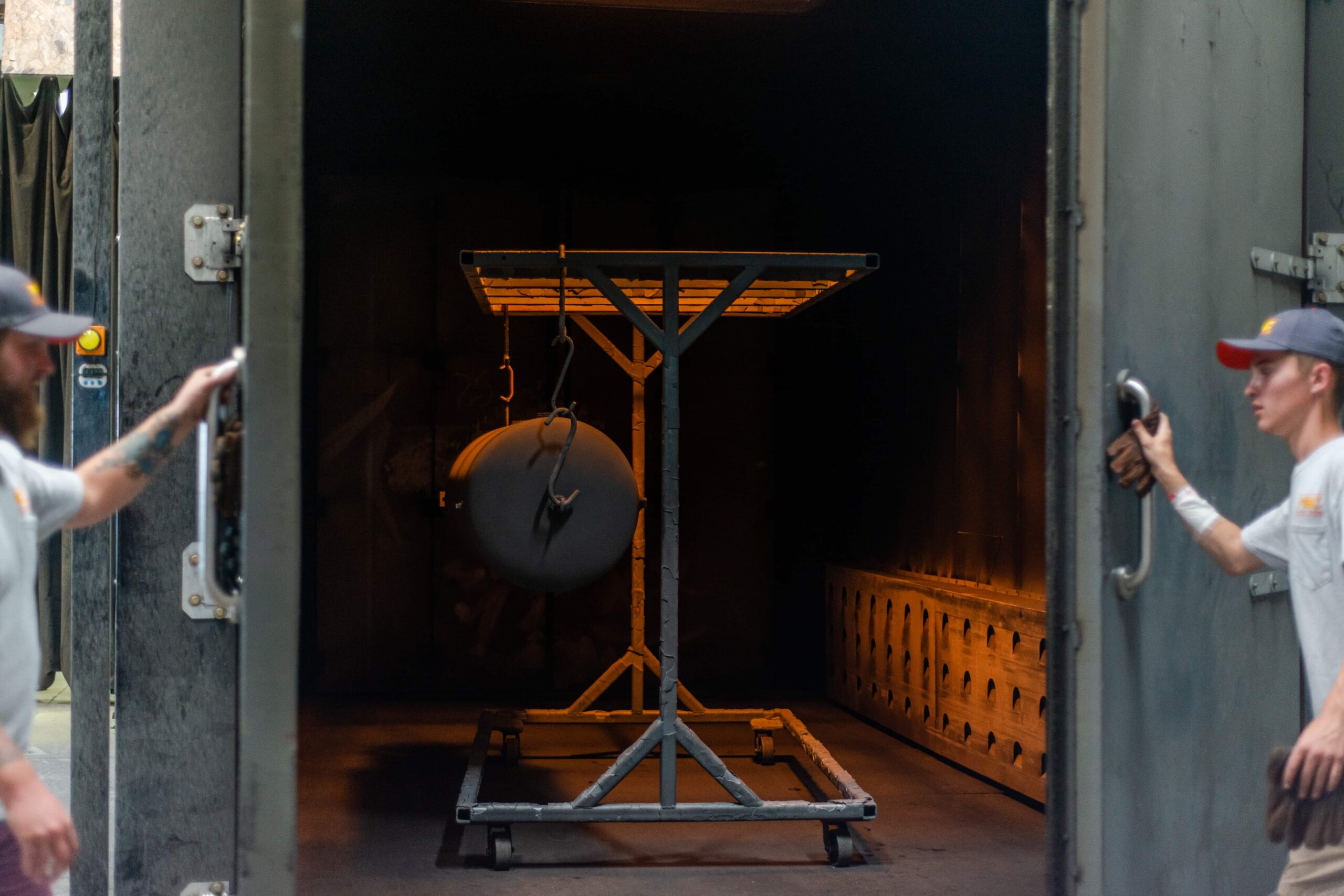 This image shows the interior of an industrial oven or kiln used for powder coating and metal resurfacing. In the center of the oven is a metal rack or stand with a circular object (likely a metal part) suspended from it, ready for treatment. The oven has a warm orange glow from heating elements, and visible perforated panels line the interior walls. Two workers in caps and light-colored shirts are positioned on either side of the open oven doors, appearing to be managing the loading or monitoring process. This industrial equipment represents a key step in the powder coating process where metal items are heated to cure the applied coating.