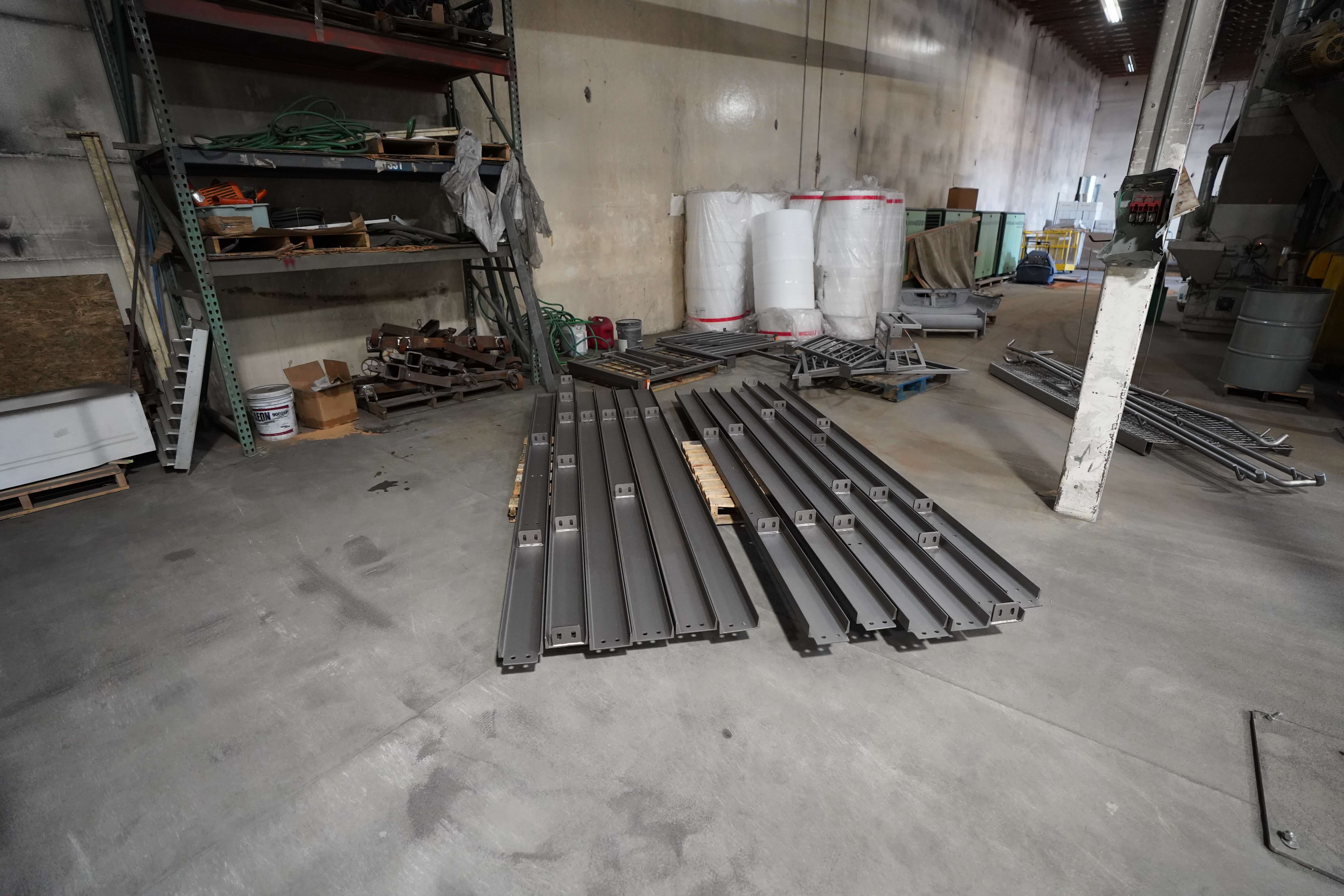 The image shows a storage or staging area of the metal fabrication facility. In the foreground are several long, dark gray metal beams or posts laid out on the concrete floor. These appear to be structural components with pre-drilled mounting holes, likely for the railing or fence systems