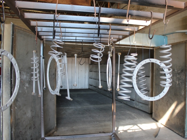 White metal springs and rings suspended in an industrial powder coating workspace, hanging from overhead hooks inside a concrete-walled preparation room with natural light filtering in