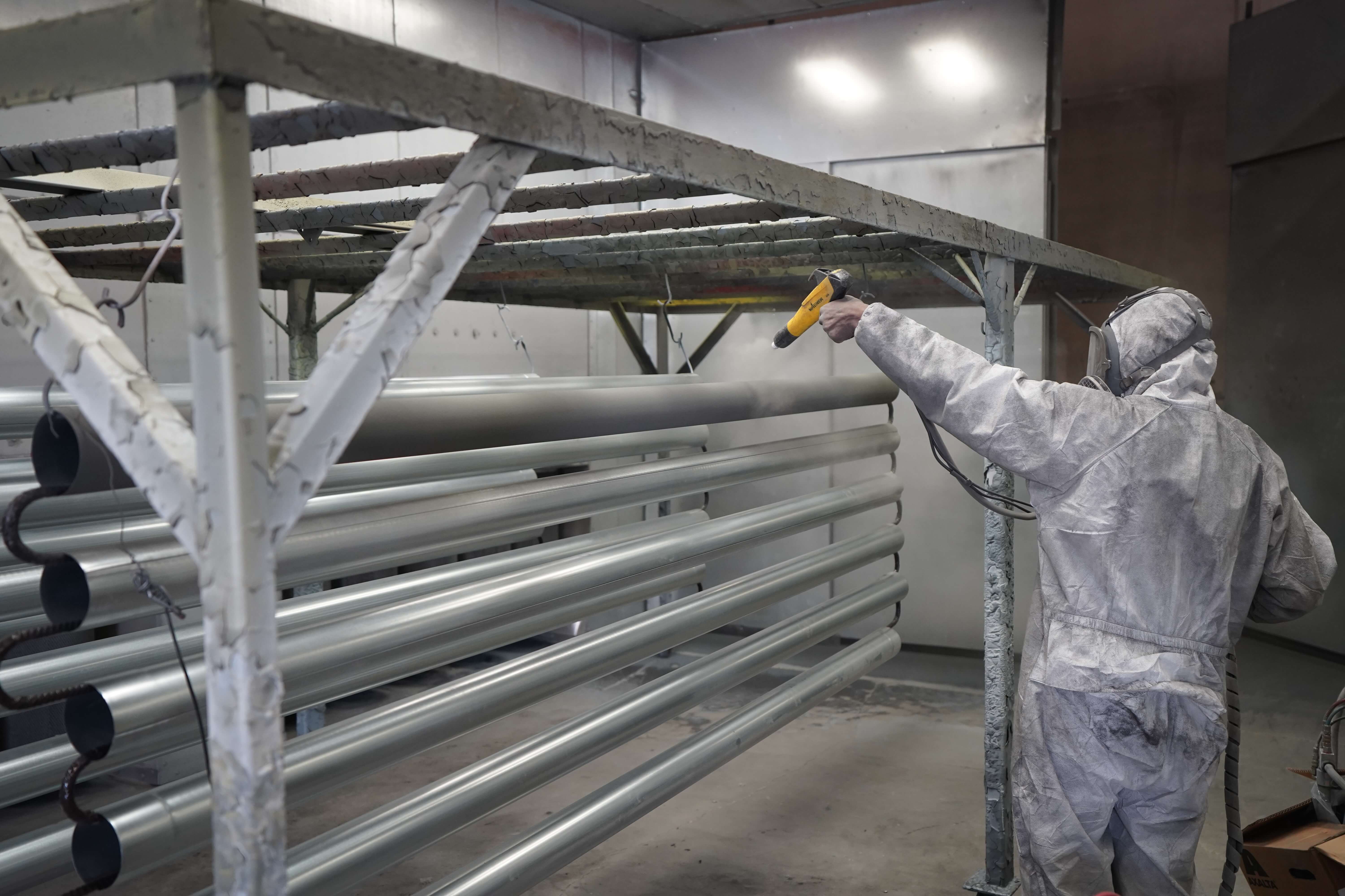 A technician wearing protective white coveralls and hood applying powder coating to aluminum pipes using a yellow electrostatic spray gun. The silver metal tubes are arranged horizontally on a specialized rack for even coating coverage in a professional powder coating spray booth