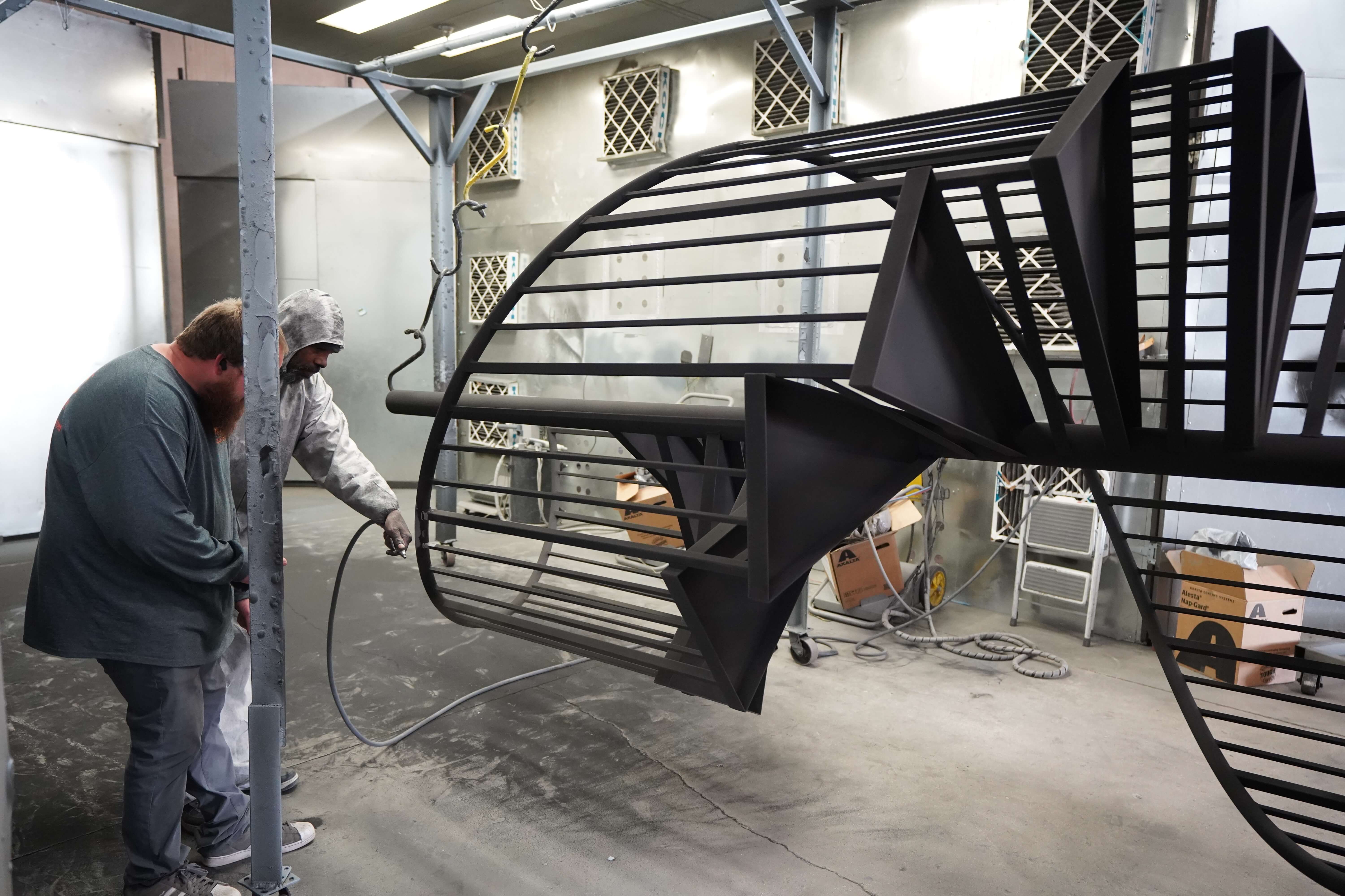 The image shows an indoor paint or powder coating area of the metal fabrication workshop. In the foreground, a curved black metal staircase or spiral staircase component is suspended from hooks or hanging apparatus, appearing to be in the process of being finished. The metal structure features horizontal railings with vertical supports