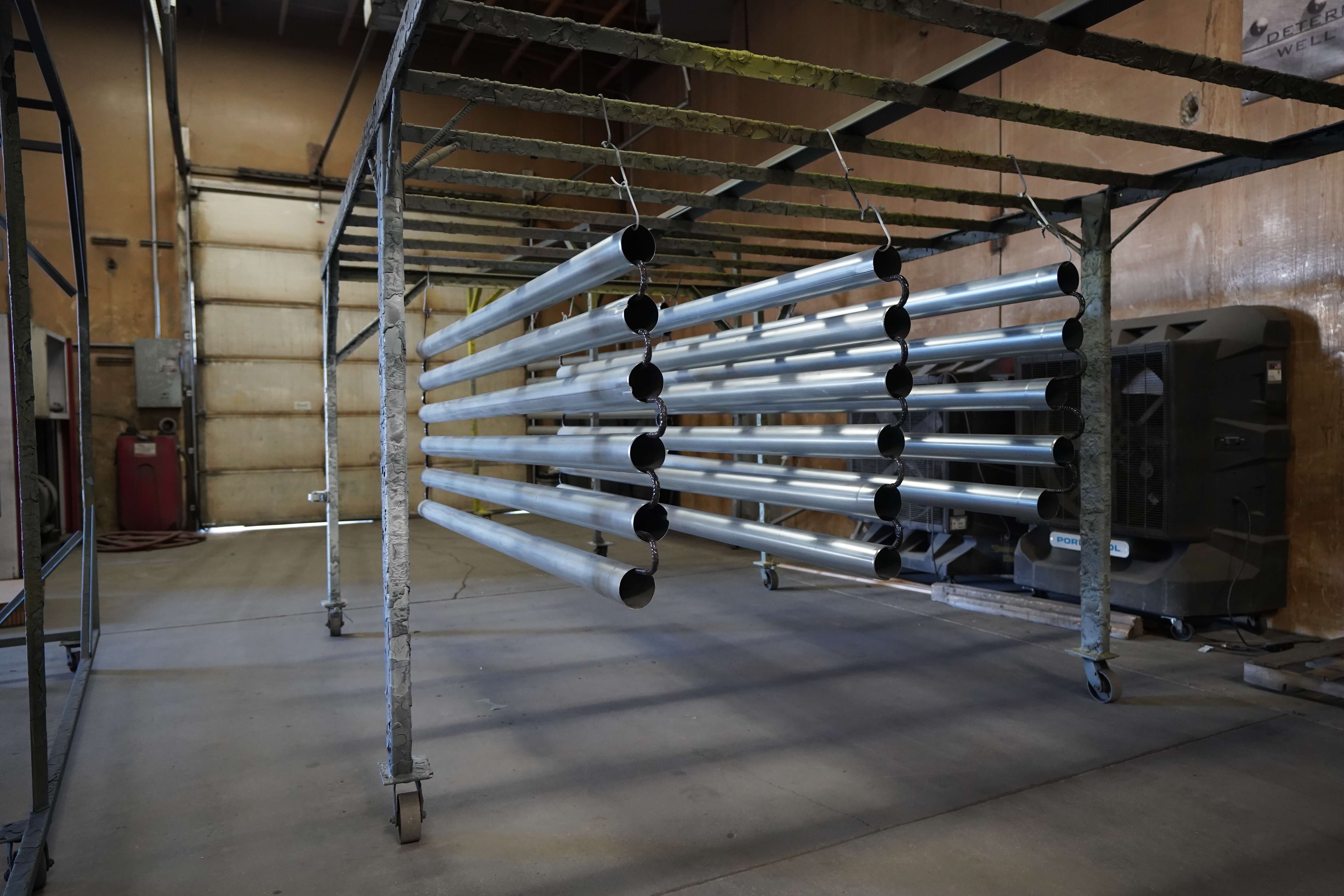 This image shows multiple aluminum or metal pipes hanging from a metal rack or drying station in what appears to be an industrial workshop or manufacturing facility. The silver cylindrical pipes are arranged horizontally in rows, suspended from hooks or chains on a movable metal frame with wheels. The pipes appear to be either freshly fabricated, in the process of being coated, or drying after treatment. The workshop has beige/tan walls, a concrete floor, and industrial equipment visible in the background, including what might be a gas tank or compressor on the left side. This appears to be part of a metal fabrication or finishing process where components are hung to dry after painting or other surface treatment.RetryClaude can make mistakes. Please double-check responses.