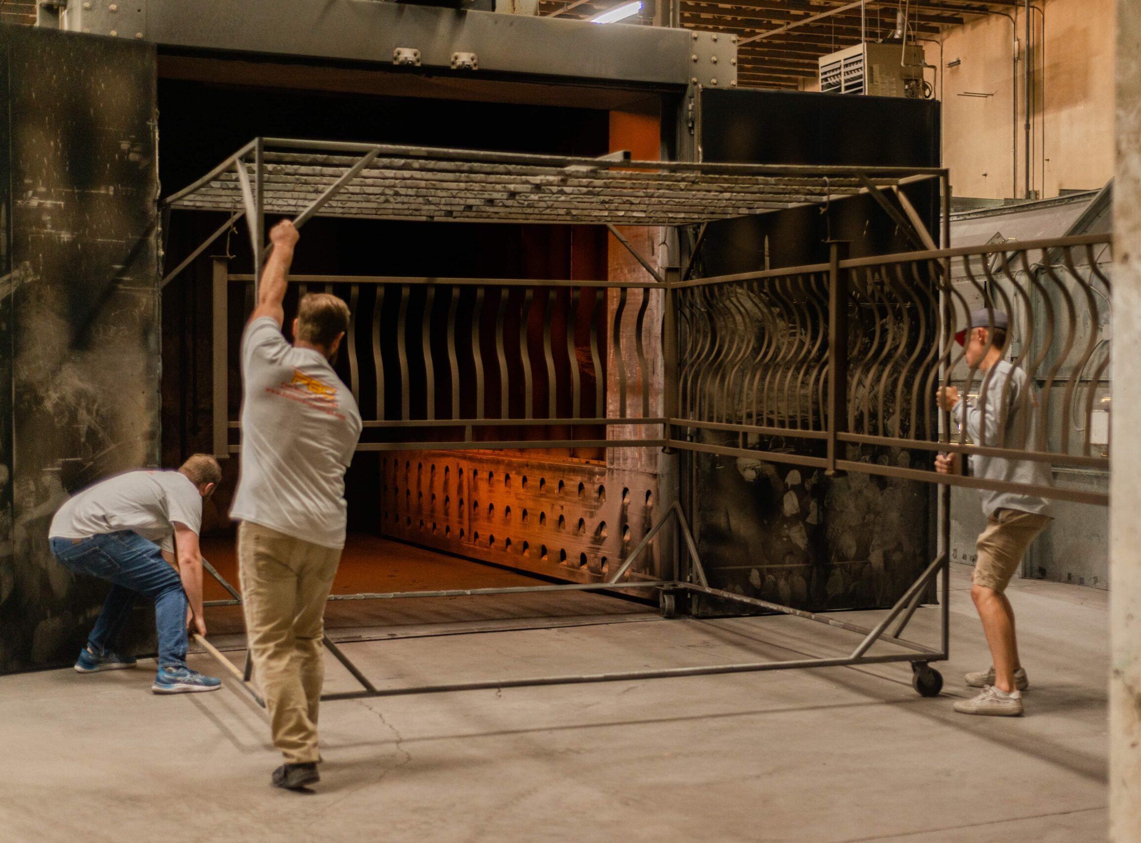 Three workers in a powder coating facility working together to maneuver a large metal rack cart. The team is positioning the cart near an industrial curing oven with glowing orange interior visible. One worker lifts the top frame while two others adjust the base. All workers wear company branded gray t-shirts, and metal railings for powder-coated items can be seen inside the oven. The cart appears to be used for transporting metal components through the powder coating process.
