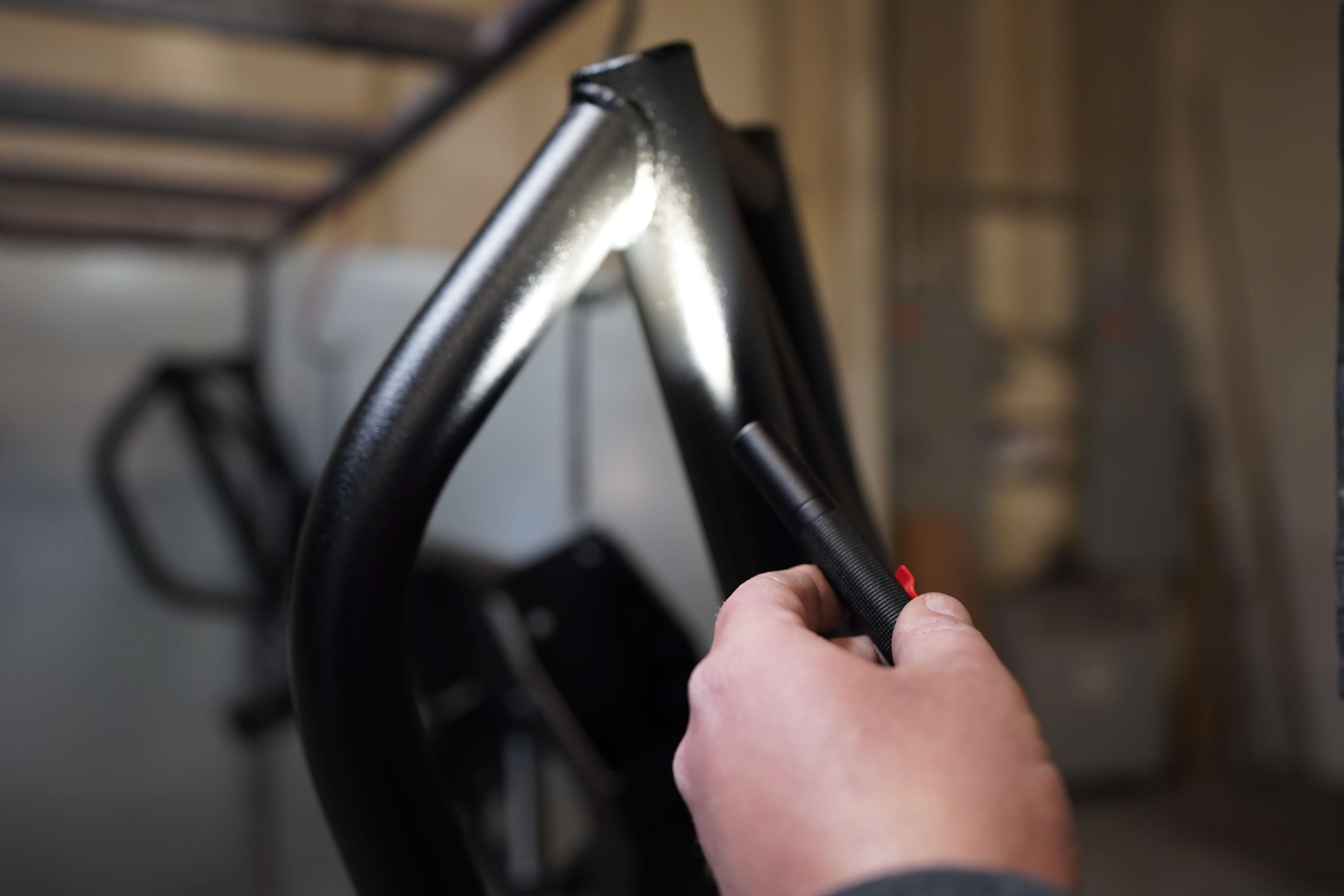 The image shows a close-up of a hand holding what appears to be part of a bicycle frame, specifically the head tube or front fork area. The frame has a sleek, glossy black finish with smooth welding at the junction points. The person's hand is gripping what looks like a handlebar or part of the frame, with a textured grip area visible. The background shows a workshop or manufacturing space that's slightly blurred, suggesting this might be in a bicycle manufacturing, assembly, or repair facility. The lighting highlights the smooth curves and metallic finish of what appears to be a high-quality bike frame component.