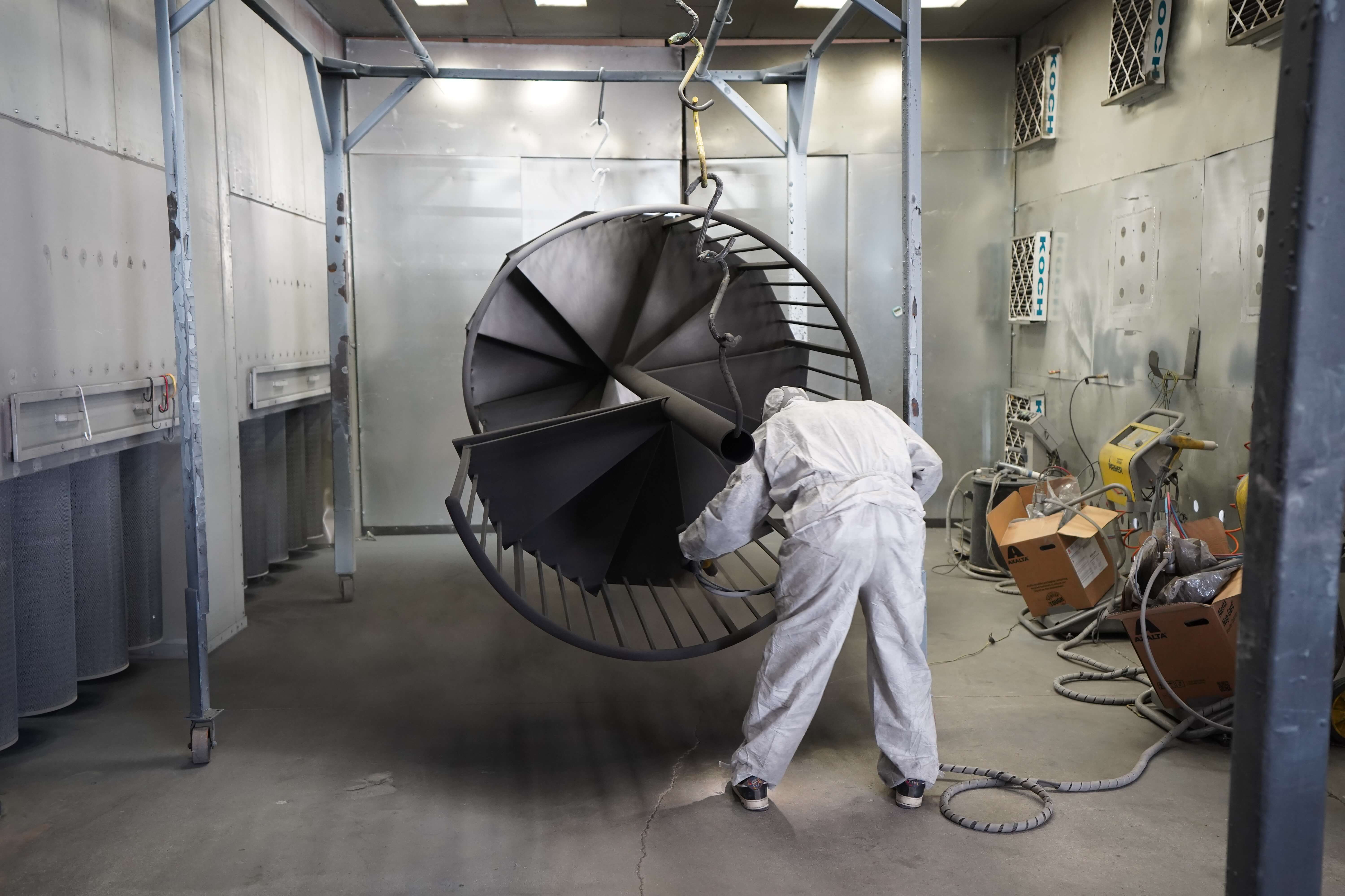 This image shows a person in a white protective suit and hood working in what appears to be an industrial spray painting or powder coating facility. They are handling a large, circular metal fixture that resembles a spiral staircase or helical structure suspended from an overhead hook. The worker is spray painting or powder coating the metal structure, which is dark in color (likely black or dark gray). The environment is a controlled spray booth with metal walls, ventilation systems, and various equipment visible in the background. This represents a typical industrial finishing process where metal components are coated for protection and appearance.