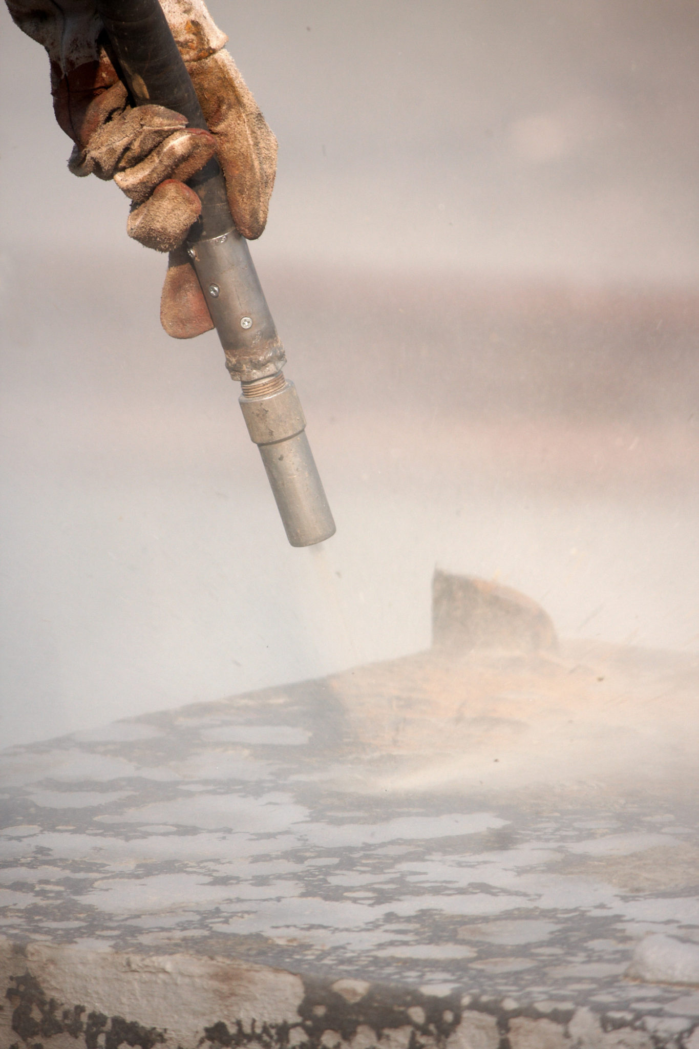 Close-up of a gloved hand holding a sandblasting or industrial cleaning nozzle, creating a misty spray over a textured concrete or stone surface