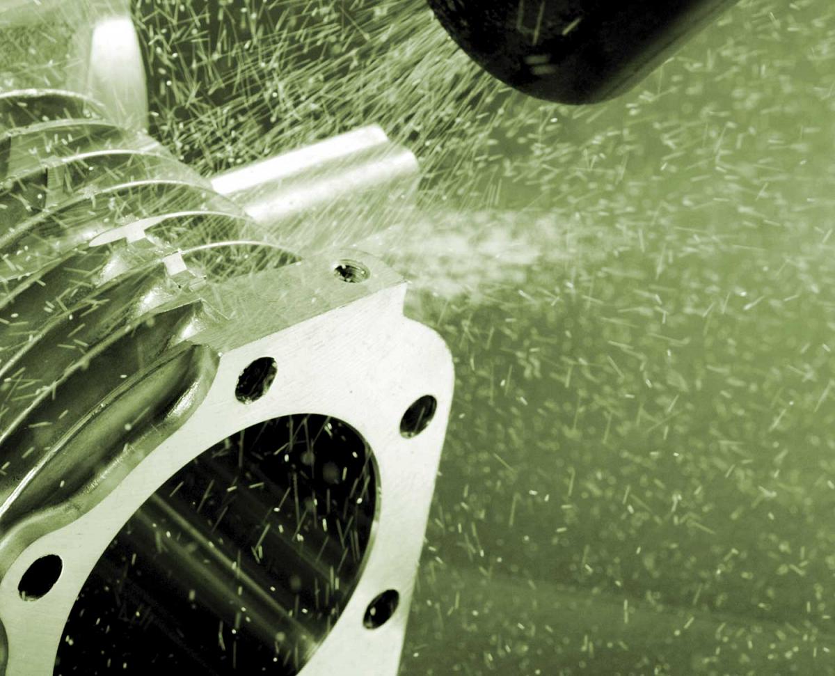 This image shows a close-up of a metalworking or machining process captured with a green-tinted filter. A metal part with mounting holes is visible in the foreground, likely a bracket or flange. Small metal shavings or debris are visible scattered throughout the image, suggesting this is capturing a drilling, milling, or cutting operation in progress. The particles appear to be flying from a machining process, creating a spray pattern across the green background. This represents precision metalworking in a manufacturing environment, where raw metal components are being shaped, drilled, or modified with industrial machinery.