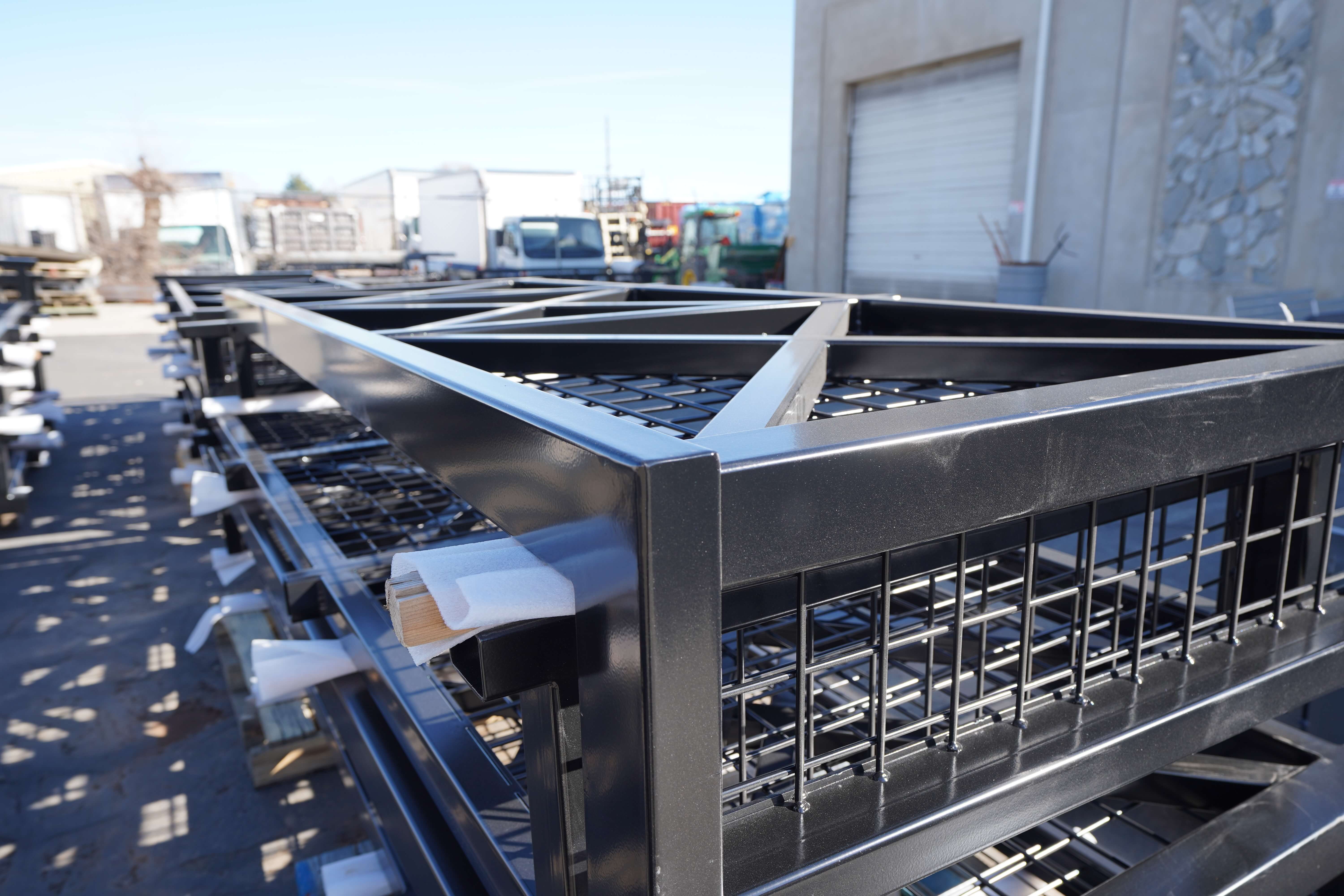 This image shows a closer view of the finished or nearly finished metal fabrication products in the yard of the metal workshop. In the foreground are several black powder-coated or painted metal frames with grid infill panels. The frames have a clean, professional finish with square corners and appear to be railing or fence sections with metal grid inserts.