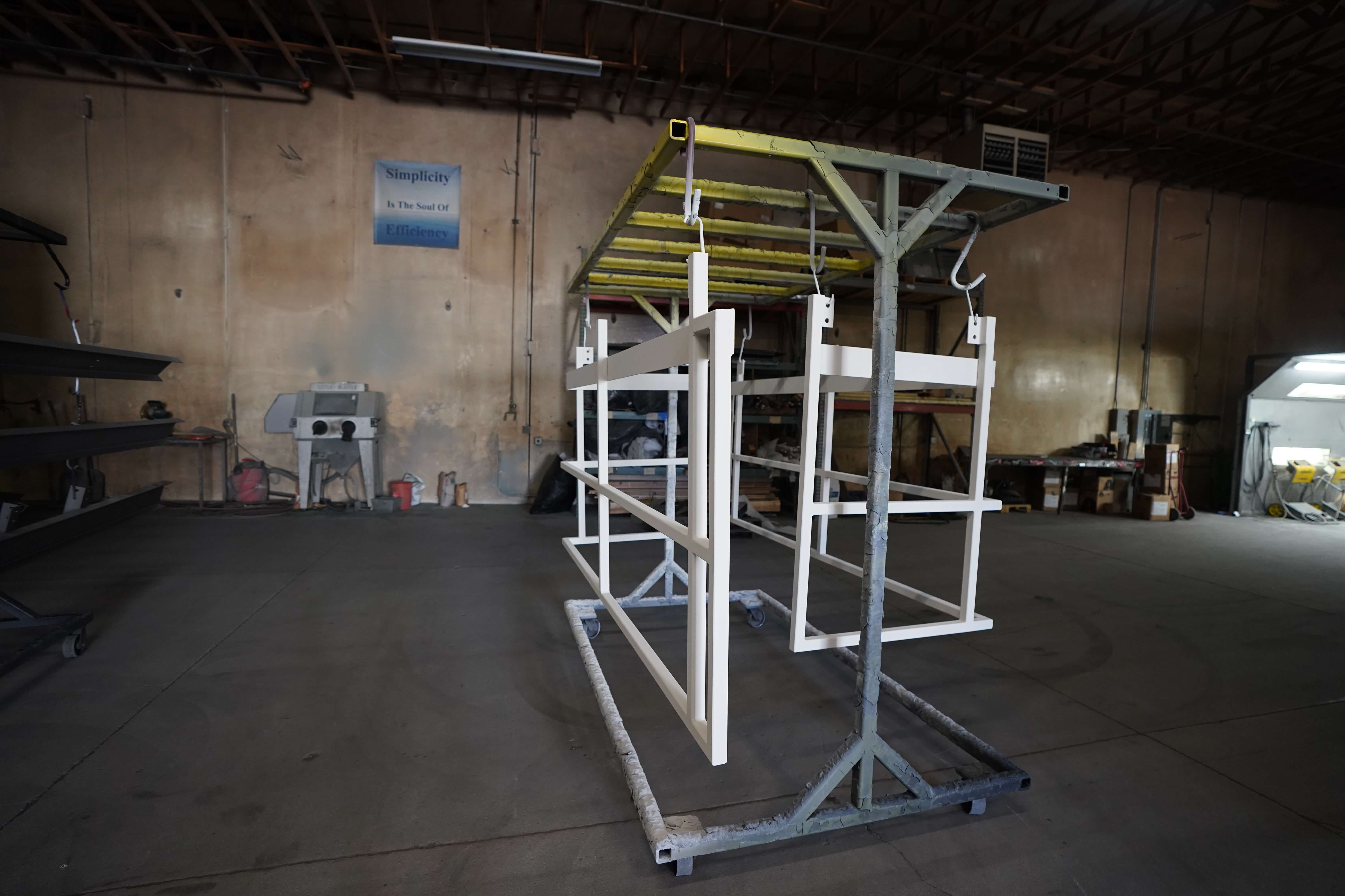 Industrial workshop interior with a metal frame structure in the center. The structure has white vertical and horizontal beams forming a rectangular framework, with a yellow-green roof section on top. The setup appears to be on wheels and is positioned on a concrete floor. In the background, a beige wall displays a sign reading 'Simplicity Is The Soul Of Efficiency,' and various workshop equipment and tools can be seen along the walls. The ceiling reveals exposed structural elements and lighting.