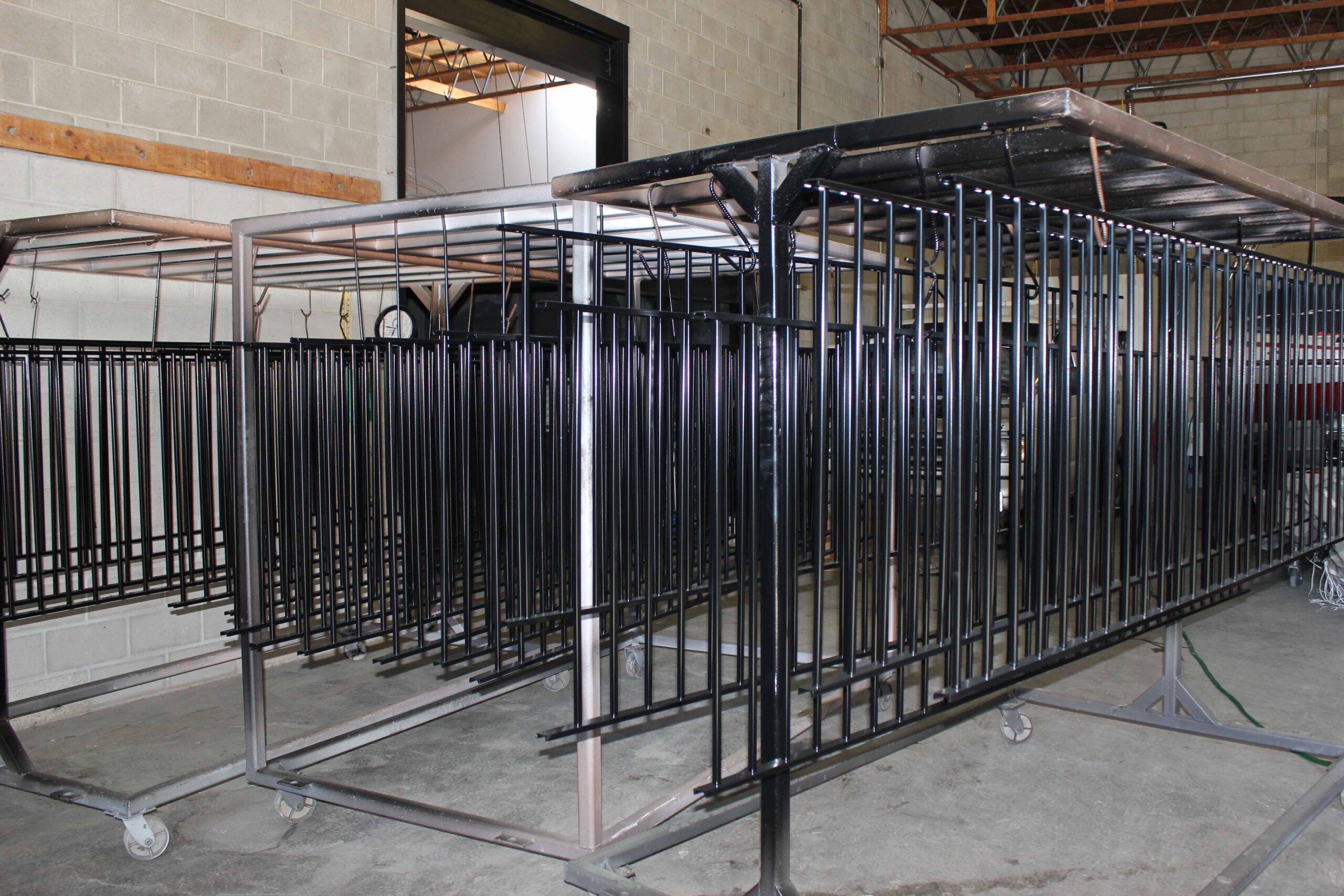 Large metal rack with vertical bars and wheels, used for powder coating or industrial part processing, situated in a concrete industrial workspace with exposed ceiling trusses