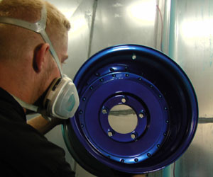 Industrial worker wearing a respirator mask inspecting a blue metal wheel or component during powder coating or metal finishing process