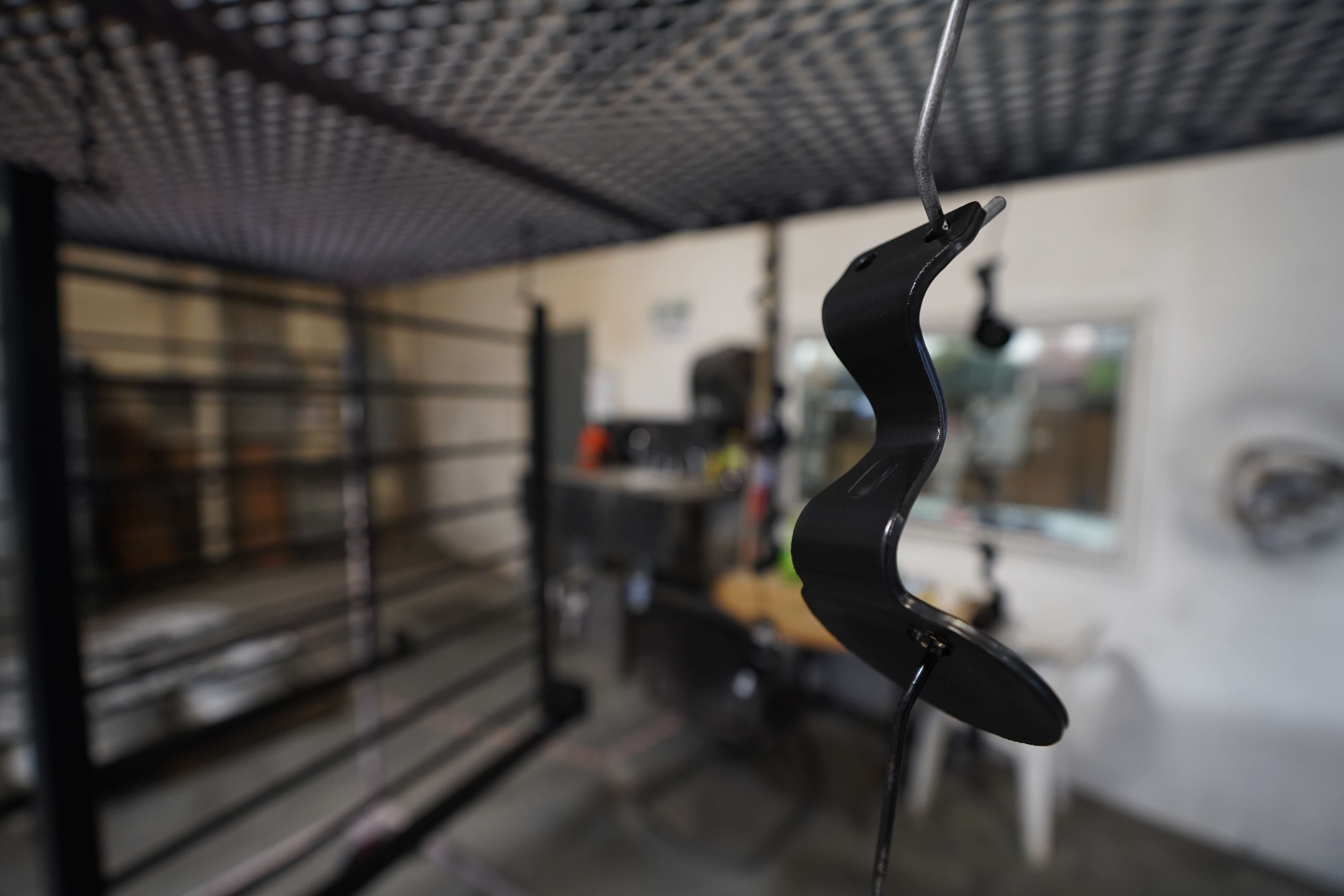 A black metal object with a curved, wavy shape hanging from a wire in what appears to be a workshop or garage. The object is in focus in the foreground, while the background shows a blurred view of equipment, shelving, and a grated ceiling structure. The space appears to be an industrial or manufacturing environment
