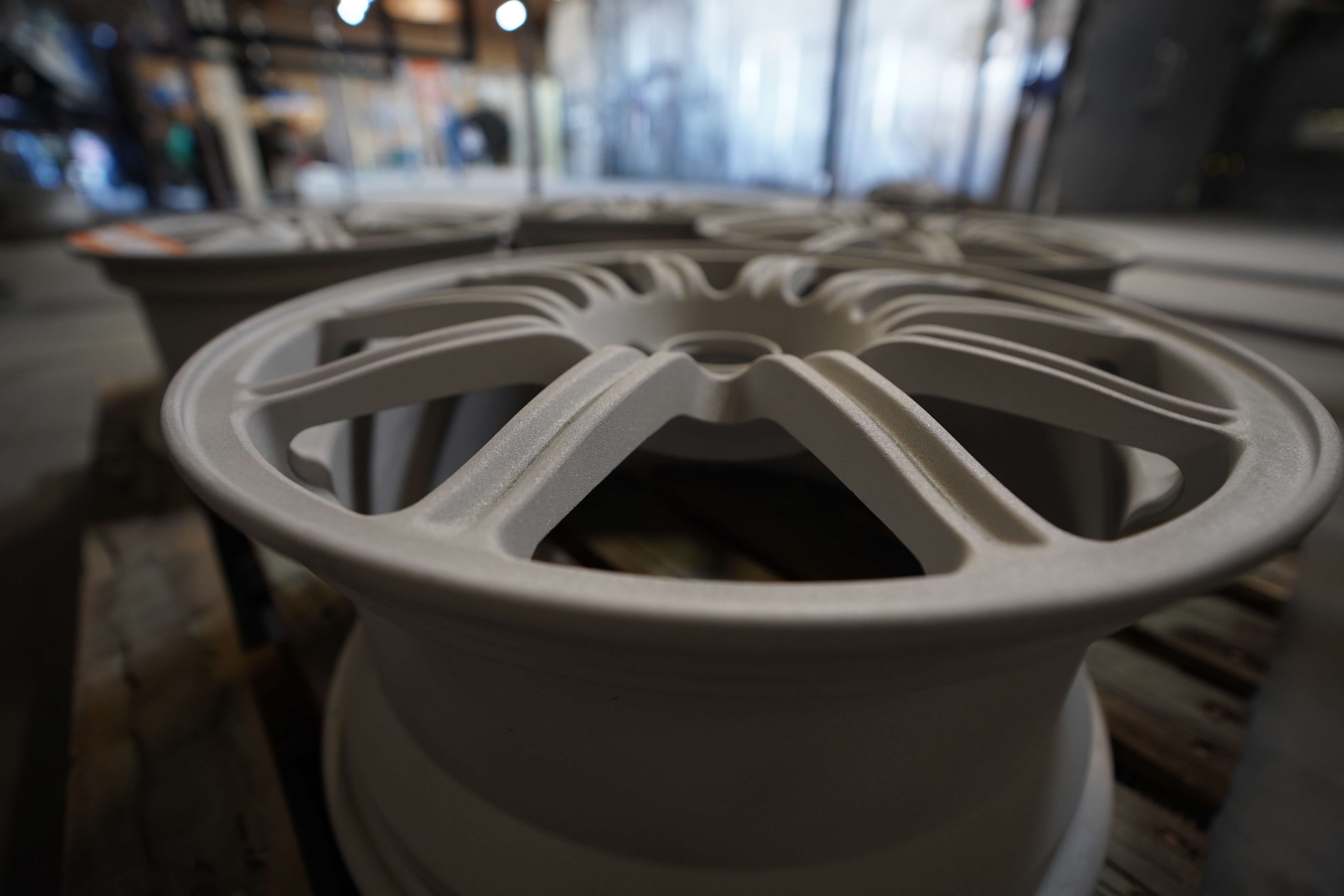 Close-up of a silver alloy wheel rim in a workshop. The wheel has a multi-spoke design with a matte finish. Several other wheels can be seen blurred in the background of what appears to be an automotive manufacturing or repair facility