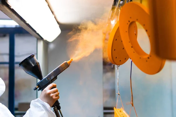 Worker applying orange powder coating to metal parts in a controlled spray booth.