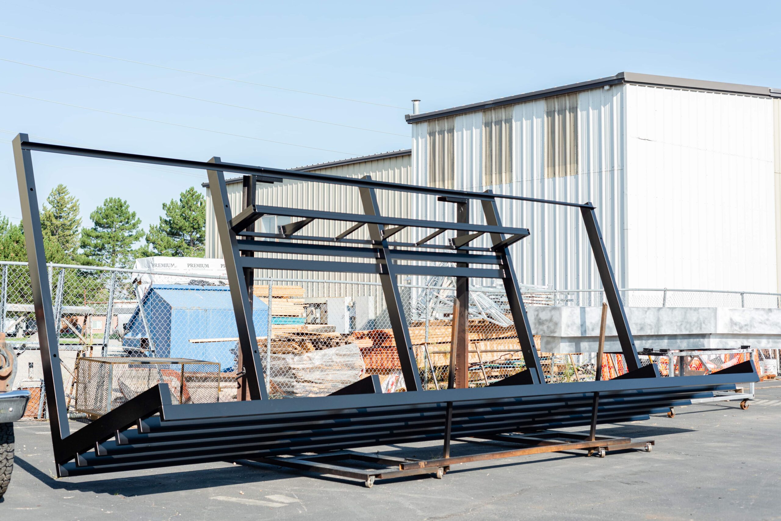 A large powder-coated metal frame structure positioned in an outdoor industrial area. The black metal framework appears to be part of a commercial sign or canopy system that has been fabricated and powder coated. The substantial steel framing is arranged on the ground in front of a metal industrial building, likely waiting for transport or installation. In the background, there's a chain-link fence surrounding a storage yard with various construction materials and equipment. This image showcases a completed industrial metal fabrication project, demonstrating the scale and durability of powder-coated structural steel components.