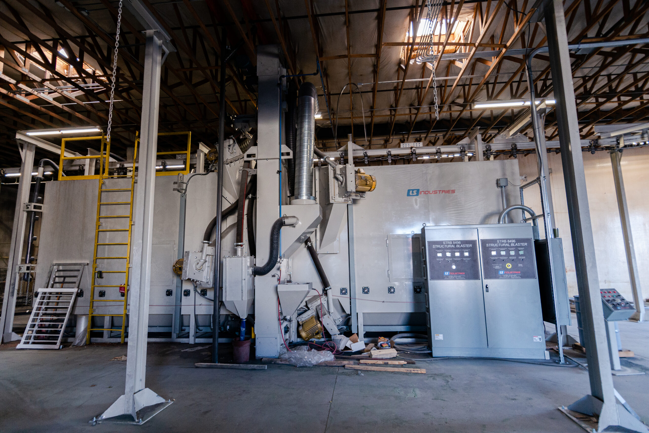 Industrial metal surface preparation equipment in a large warehouse facility. The image shows a substantial sandblasting or shot blasting system from LS Industries with "STRUCTURAL BLASTER" control panels visible. The setup includes a large gray cabinet-style machine with ventilation ducts, electrical controls, and a yellow access ladder. The equipment is housed in a high-ceiling industrial building with exposed wooden roof trusses and overhead lighting. This machinery is used for cleaning and preparing metal surfaces before powder coating application.