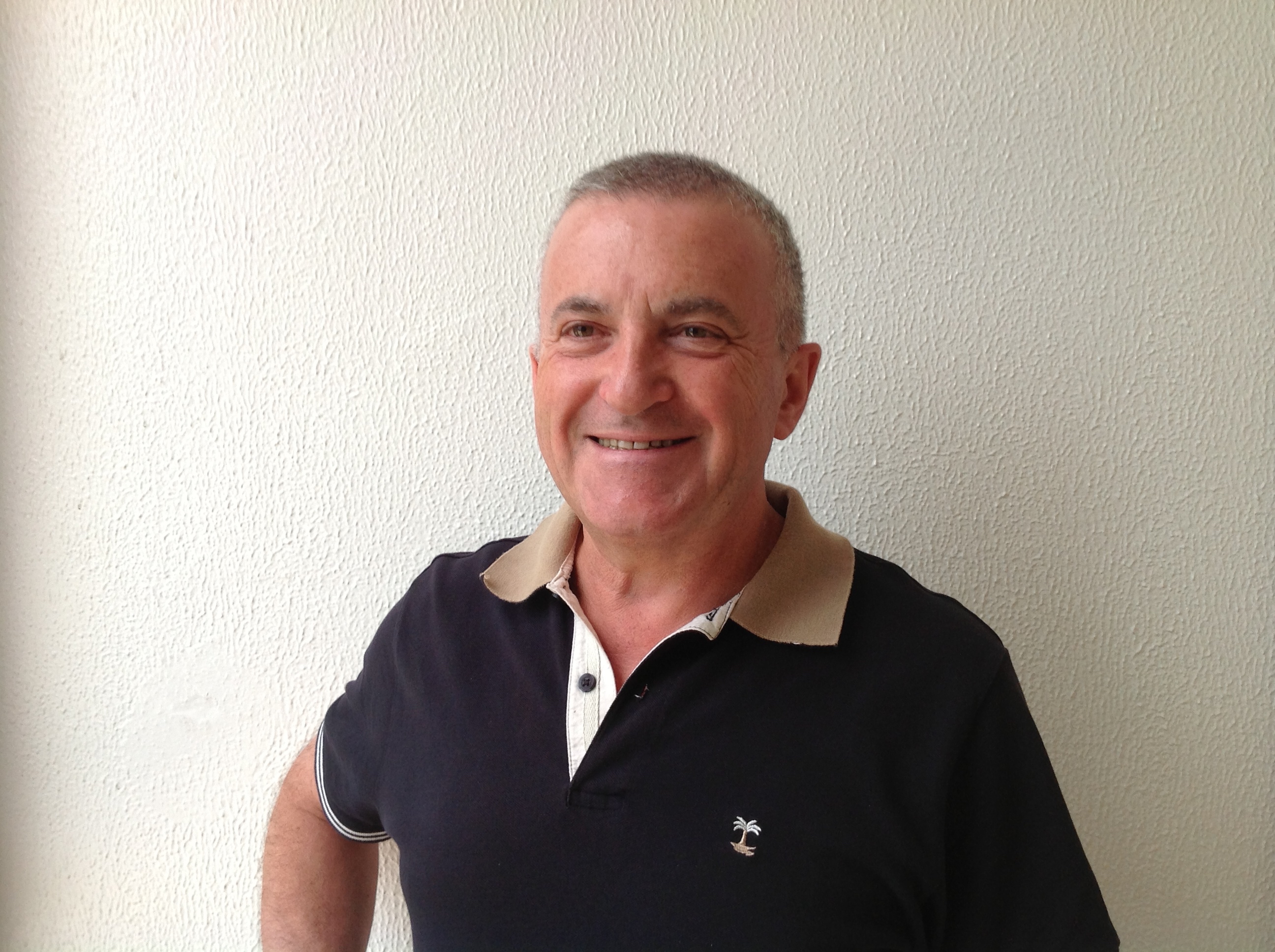 Un uomo che indossa una polo nera sorride e sta in piedi davanti a un muro bianco.