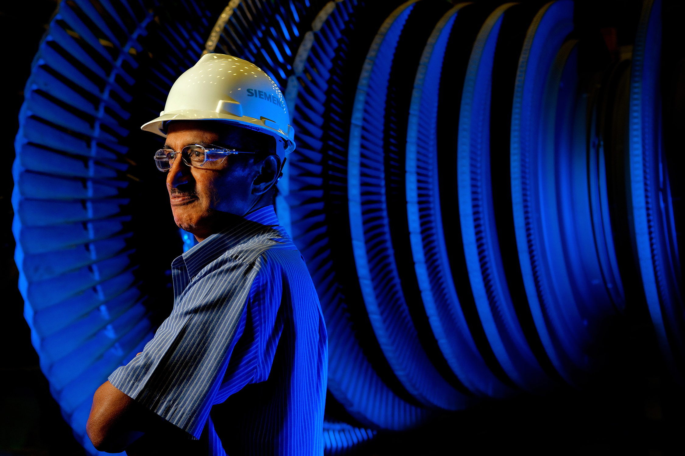 Photo is an environmental portrait of a worker in an aerospace manufacturing facility or plant. The male worker is posing for the photo. He is standing to the side of a giant turbine engine and his arms appear to be crossed. He is looking off the left side of the image. The worker, who is nicely dressed and might be an engineer, is wearing proper safety equipment, including a hard hat and safety glasses.
