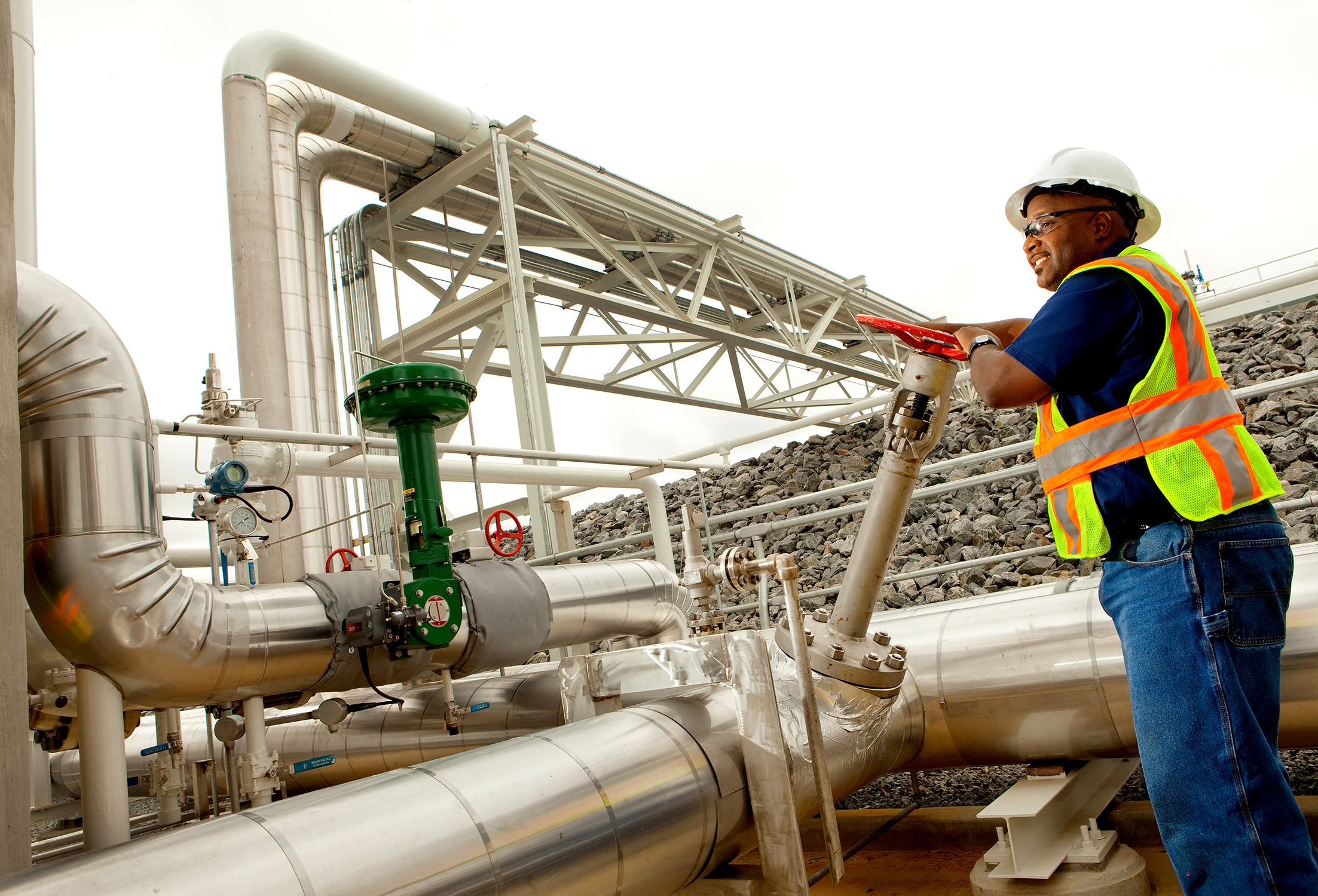 An employee at a natural gas facility smiles as she turns the valve on a pipeline. The woman was photographed using a wide angle lens to show the twists and turns of the pipeline. She is small on the right side of the image.