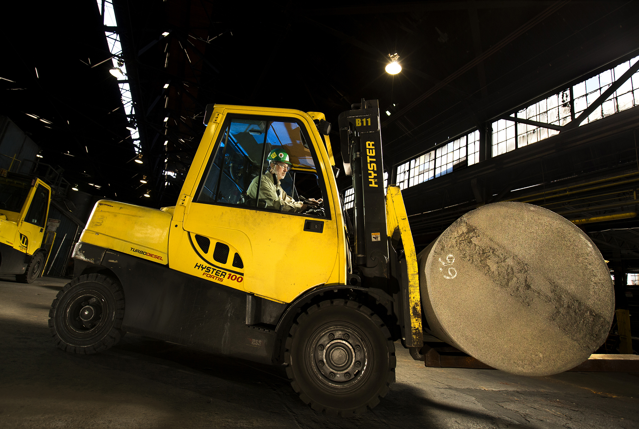 A driver uses a Hyster mover to life a large barrel in a manufacturing facility. The driver is lit using strobes so his face can be seen. The worker is wearing protective safety equipment.