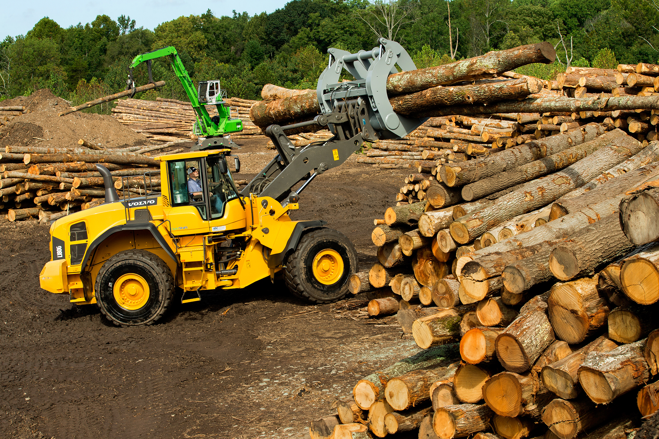Photo of two pieces of heavy equipment being used to move logs at a mill. The machine in the front of the photo has a grip that is picking up logs. It has about four logs in its teeth and is lifting them about eight feet off the ground.
