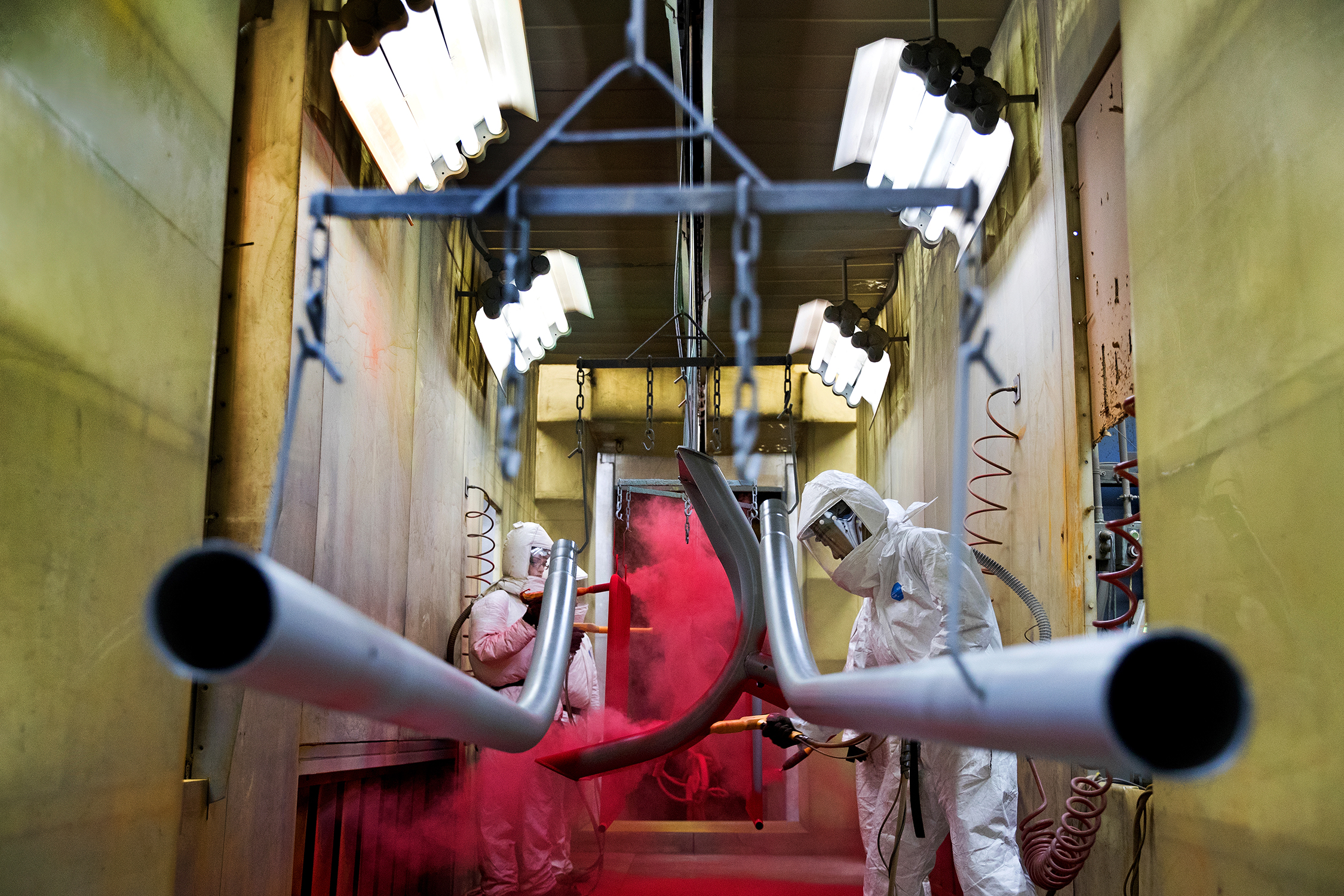 Two workers paint components in a manufacturing and assembly plant. The workers are fully clad in protective suits that have respirators connected. The suits are white and the workers are spraying red paint onto an item that is hanging between the two workers.