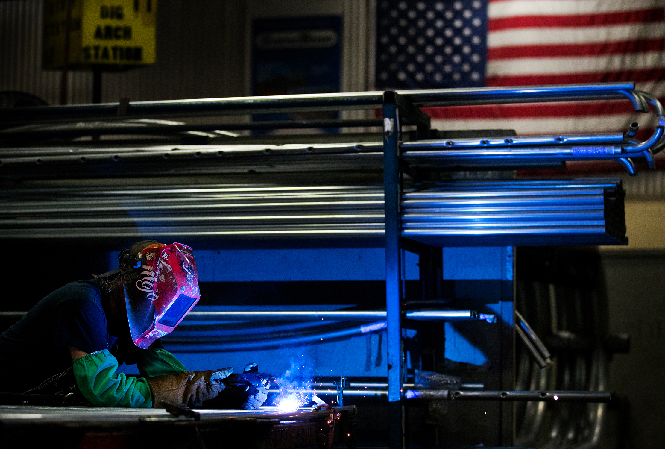 A welder is piecing together items in a manufacturing plant. The image is taken from a distance and is meant to show much of the facility. The worker is small and one the lower left corner of the photo. The worker is actively welding, and is wearing protective equipment. An American flag hangs on the wall in the upper right corner of the photo. The welding is casting an interesting blue light into the facility.