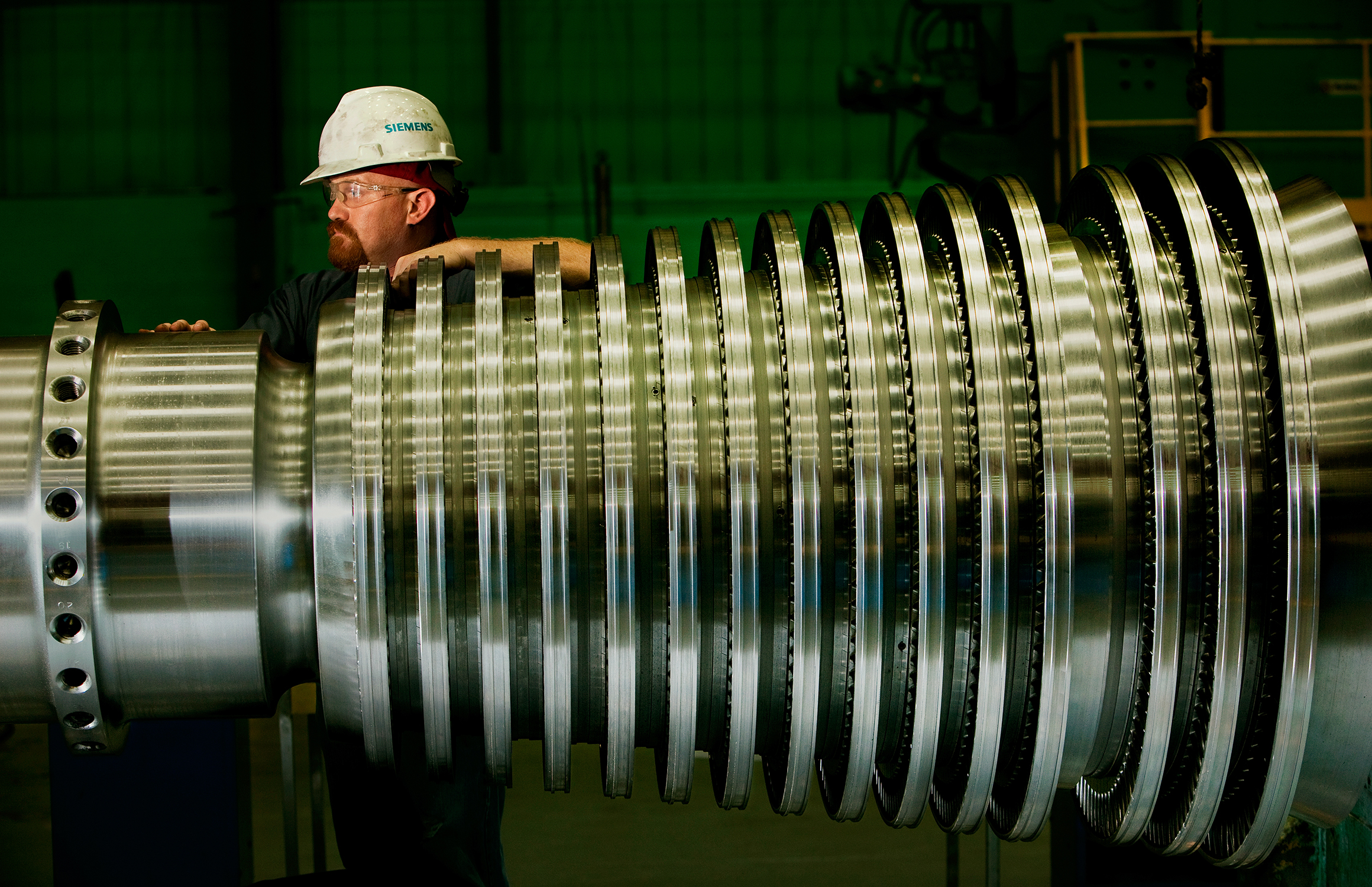 A male worker poses for the photo with his arm resting on an aerospace component, possibly part of an engine of a plane that is under construction in a manufacturing facility. The worker is looking off camera to the left. He is wearing proper safety equipment, including a hard hat and safety glasses.