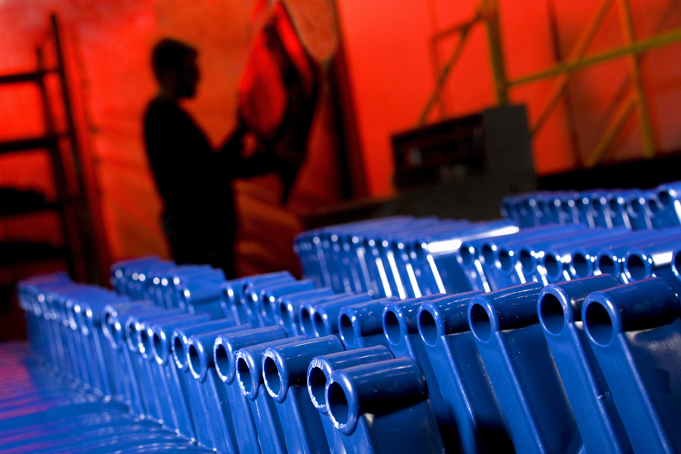 A silhouetted employee works in the background at a plastics manufacturing company. The employee appears to be looking at instructions or blueprints. In the foreground are blue plastic pieces that will make up a garbage can after they are completed.