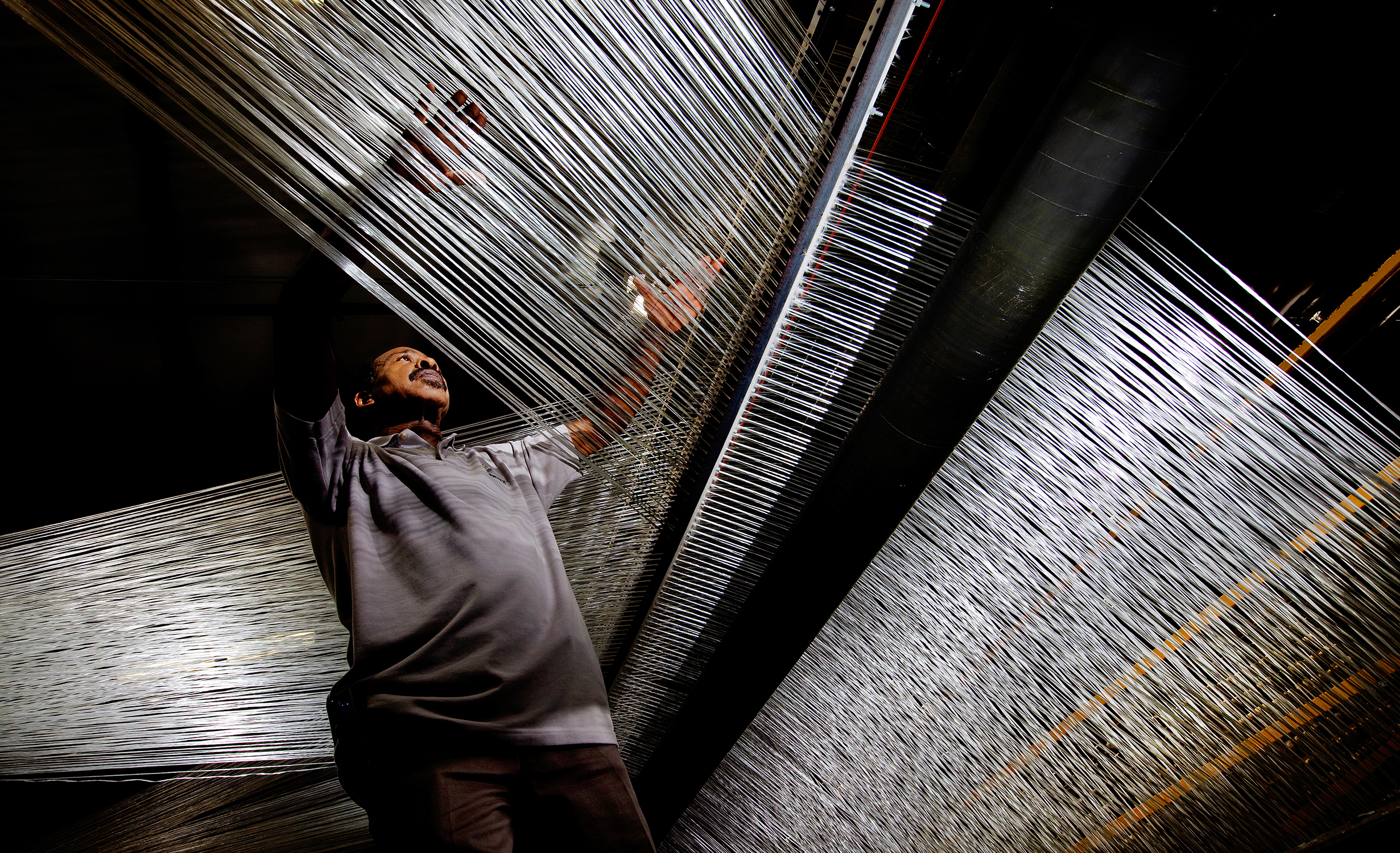 A man working in a fiber optics manufacturing facility inspects the fiber optic lines. The photo is taken from below and is looking up at the man and the underneath section of the fiber optic lines.