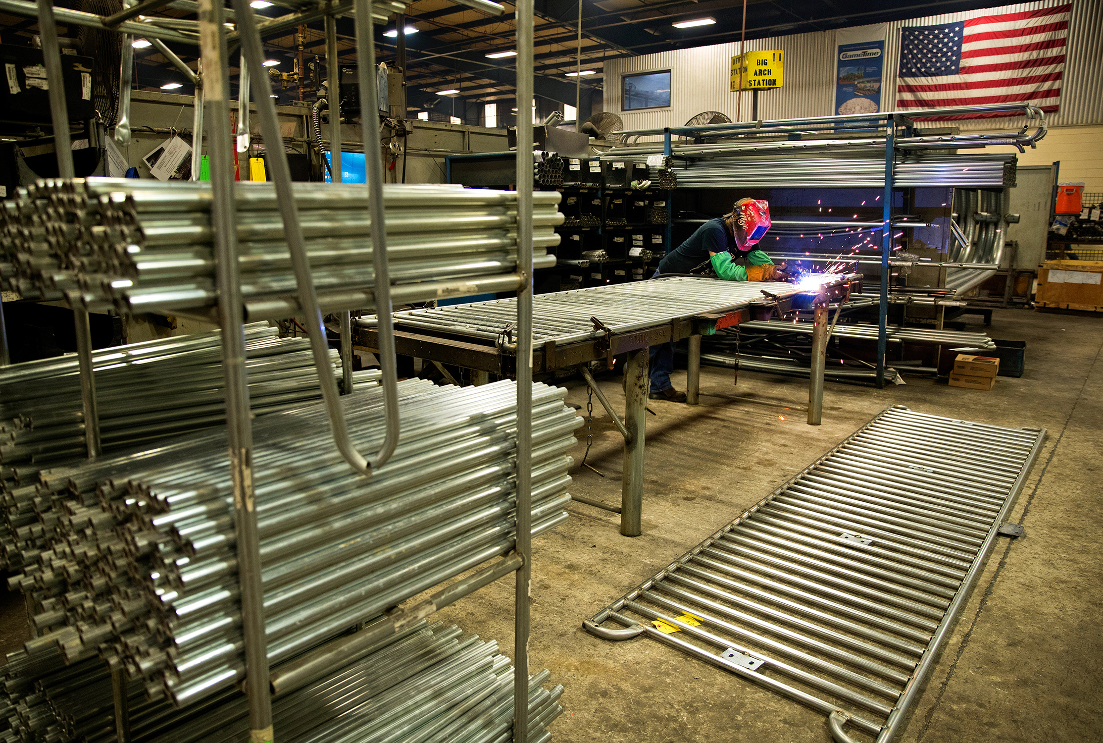 Photo of a welder in the process of welding metal in a machine shop. The welding process is kicking off sparks, and the welder is lite with the glow of the torch. The photo is taken using a wide angle lense and the welder is in the distance. The welder is wearing proper protective safety equipment.