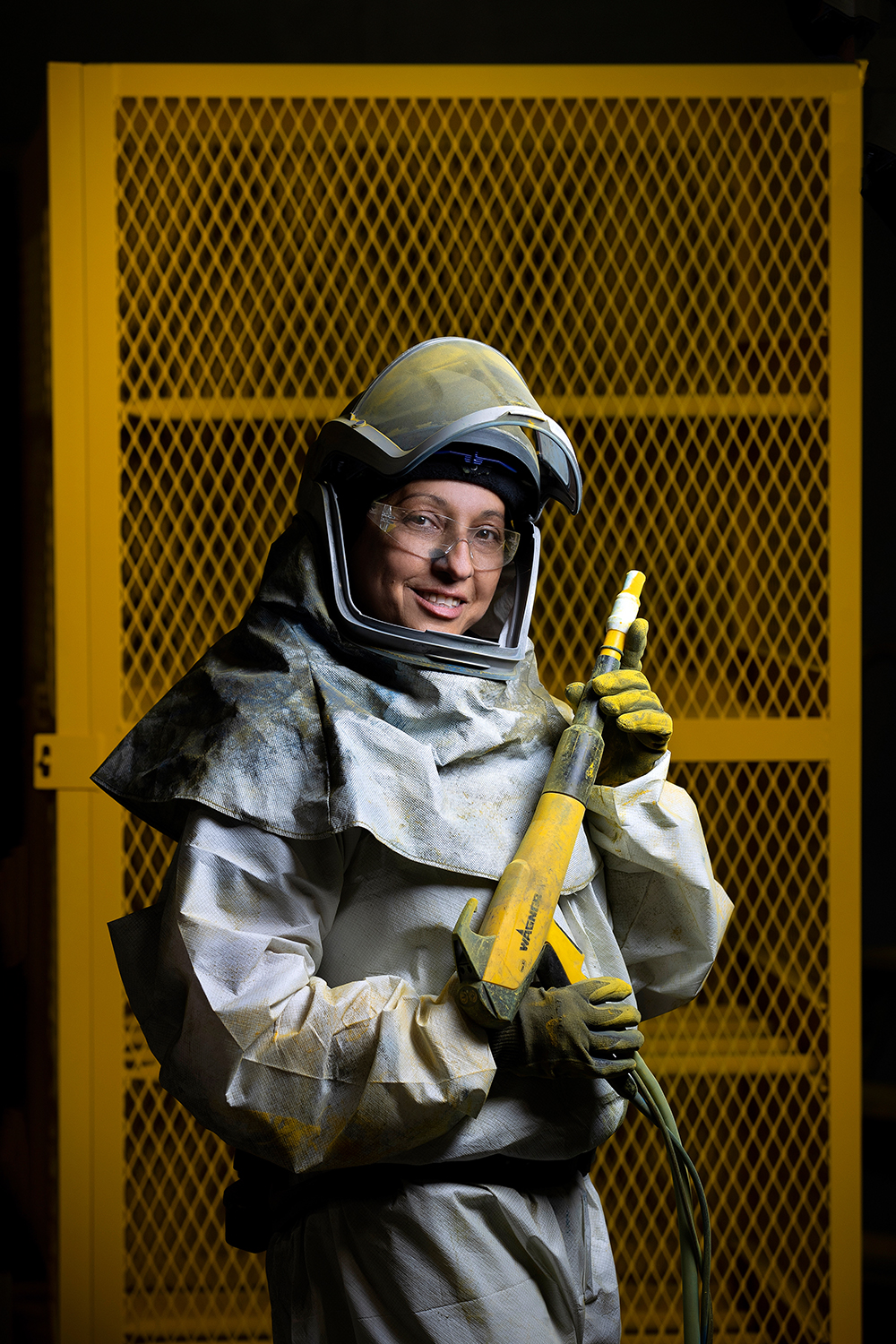 A female worker wearing a full-body safety suit poses for the camera. She is smiling. She appears to be holding a paint spray gun or painting tool and is standing in front of a large metal grate, which is painted safety yellow. The photo is an environmental portrait of the female worker in the industrial setting.