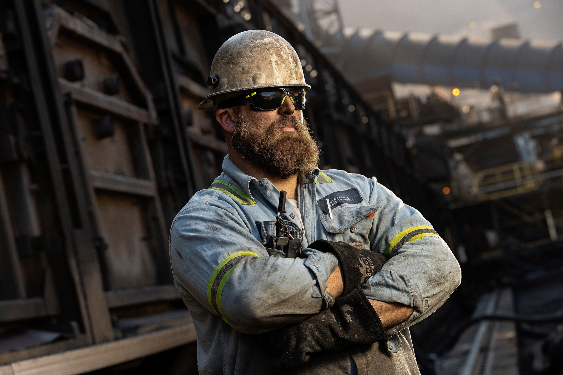 Photograph of a worker in a heavy industrial facility. Worker is posing for the camera with arms crossed. Attire is dirty and worker is wearing a hard hat, safety glasses, and heavy gloves, and appears skilled in being able to work in hard conditions like an industrial plant.